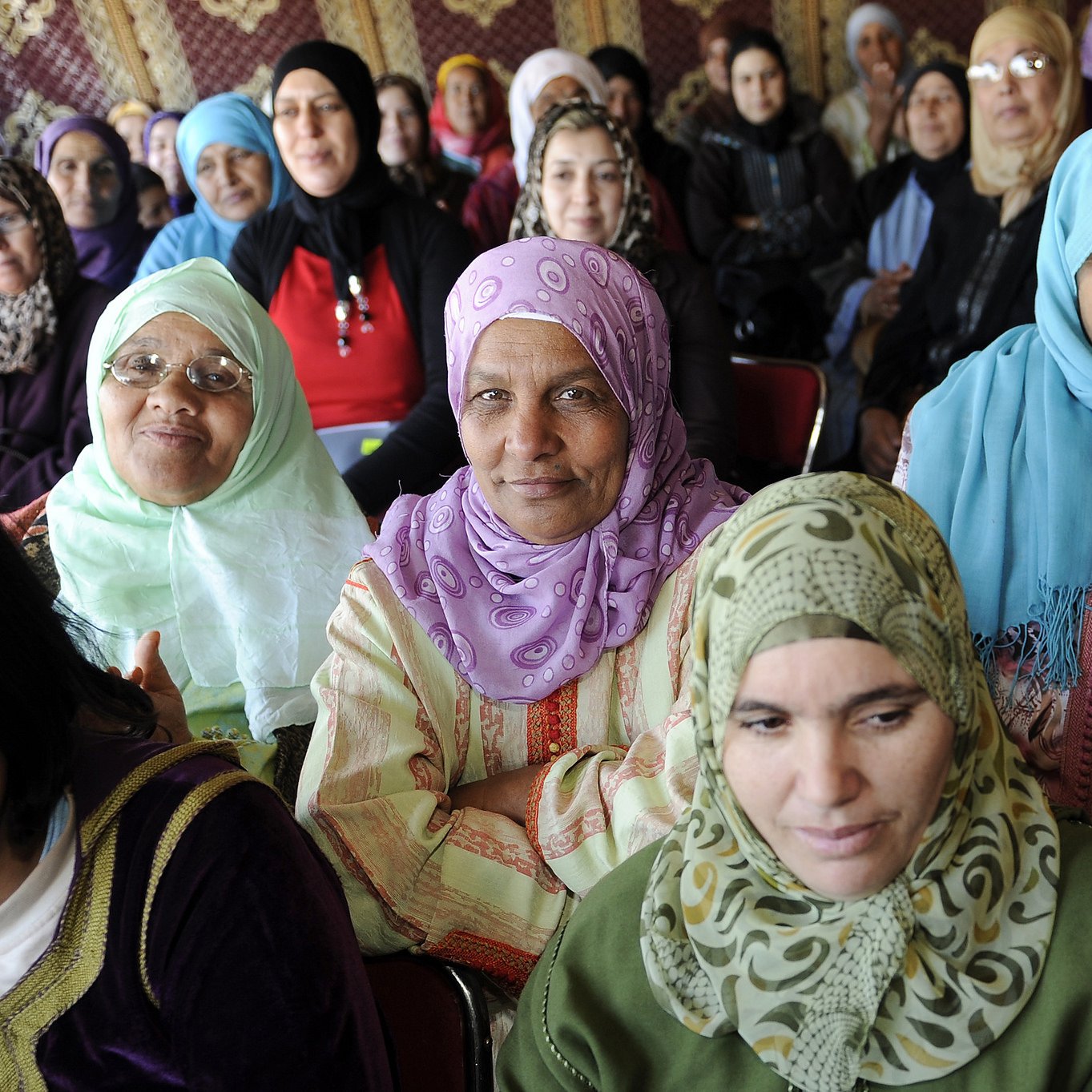 La imagen muestra a un grupo de mujeres, la mayoría de ellas vestidas con hijabs de diferentes colores y estilos. Ellas están sentadas en un ambiente que parece ser un evento o reunión, y sus expresiones varían entre la seriedad y la atención. Algunas mujeres llevan prendas tradicionales, y el fondo sugiere un entorno decorado con motivos culturales. La diversidad en los colores de los pañuelos y vestimenta resalta un sentido de unidad y diversidad entre ellas.
