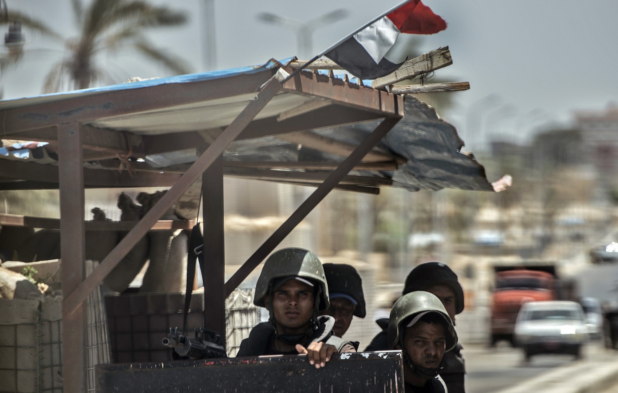الصورة تُظهر مجموعة من الجنود أو رجال الأمن، يرتدون بدلات عسكرية وخوذ، ويبدو أنهم على أهبة الاستعداد. يقفون خلف حاجز أو نقطة تفتيش، بينما يُمكن رؤية العلم المصري يُرفرف في الخلفية. المشهد يتضمن أيضًا بعض التفاصيل عن البيئة المحيطة، مثل الشوارع والمباني. الأجواء تبدو مشحونة، مما يوحي بوجود حالة تأهب أو تواجد أمني مكثف.