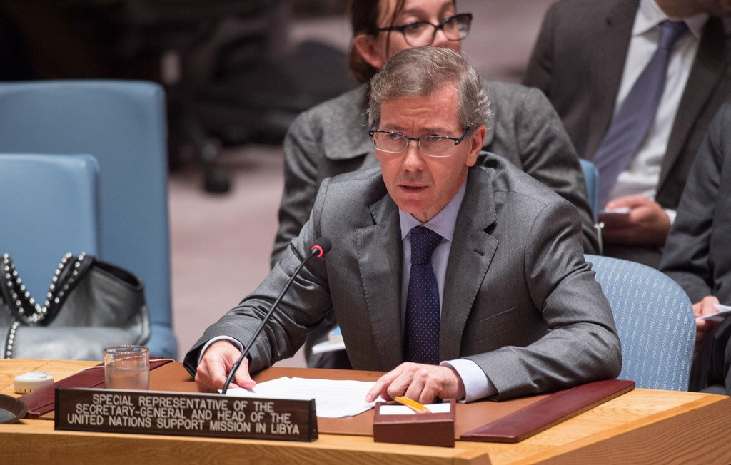 L'image montre un homme assis derrière un bureau dans une salle de réunion. Il porte un costume gris et des lunettes. Il semble s'exprimer, avec des documents devant lui. À l'arrière-plan, on aperçoit d'autres personnes, dont certaines sont en train de l'écouter. Au premier plan, un panneau indique qu'il est le représentant spécial du Secrétaire général et le chef de la mission d'appui des Nations Unies en Libye.
