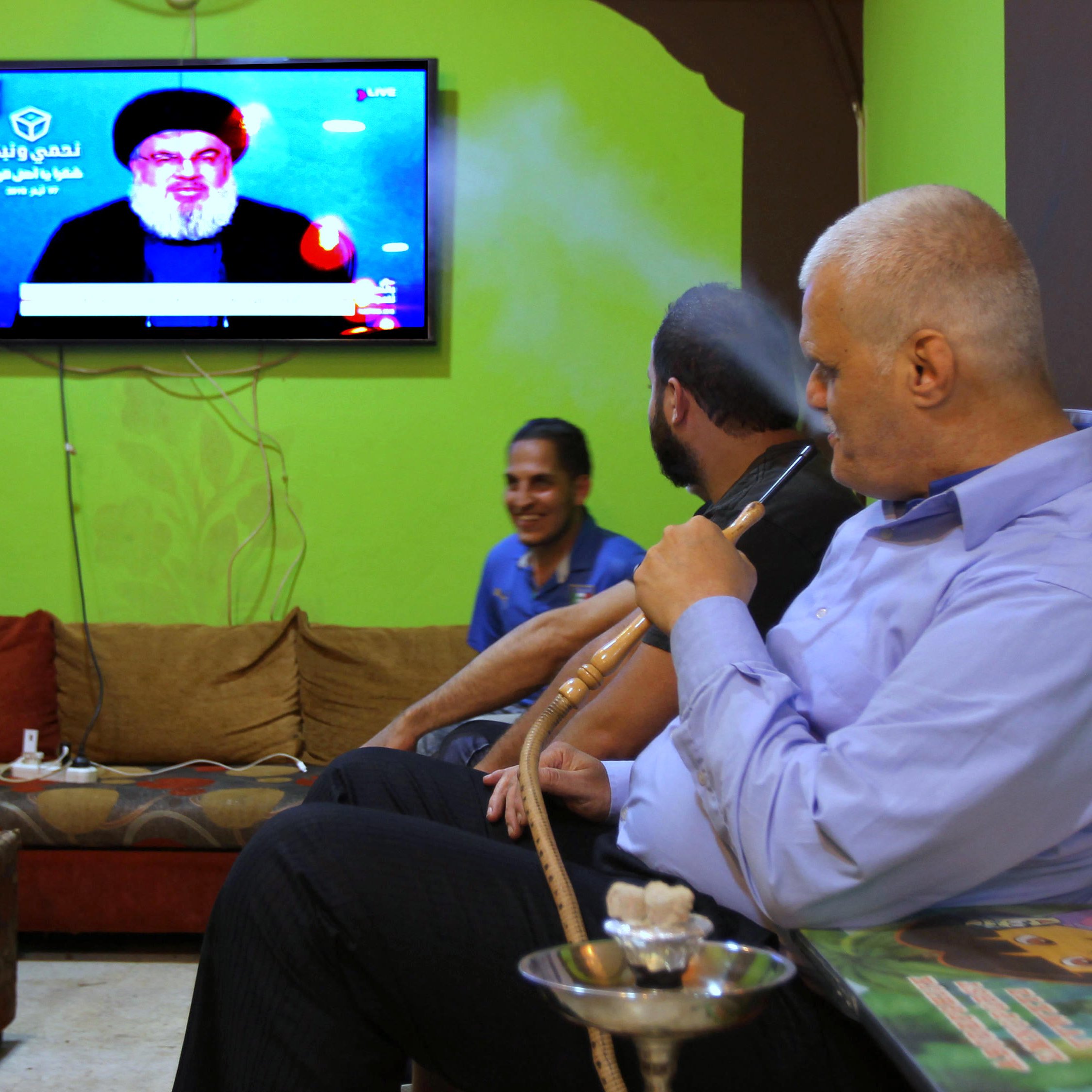 L'image montre un groupe d'hommes assis dans un salon avec des murs verts. Ils semblent regarder la télévision, où l'on peut voir un personnage à l'écran. L'un des hommes fume une pipe à eau, tandis que les autres sont engagés dans la conversation ou en observant l'écran. L'environnement est décontracté, avec des canapés confortables et des éléments de décoration simples.
