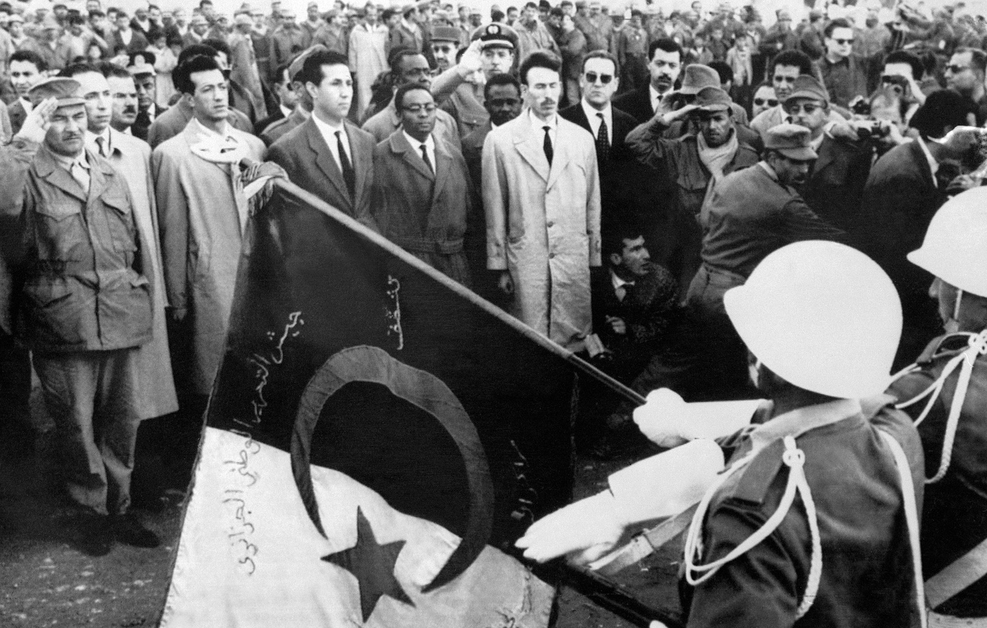 Cette image en noir et blanc semble représenter un événement historique, probablement lié à la décolonisation ou à un moment significatif dans l'histoire de l'Algérie. On peut voir un groupe de personnes rassemblées, avec des hommes en tenue civile et militaire. Au premier plan, des personnages saluent tandis que des soldats présentent un drapeau. L'atmosphère semble chargée d'émotion et de solennité, indiquant un moment important qui pourrait symboliser la fierté nationale ou la lutte pour l'indépendance.