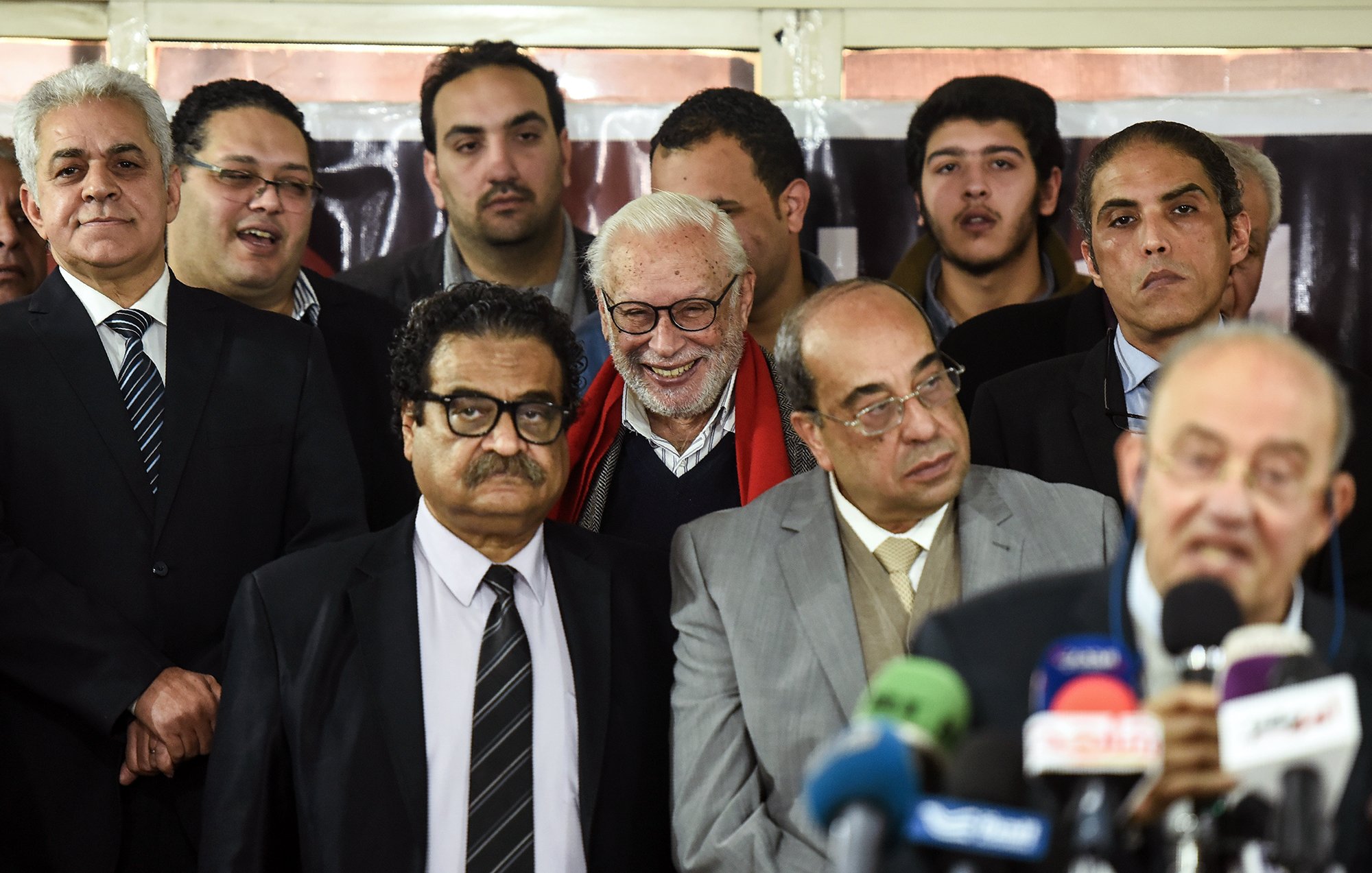 این تصویر گروهی از مردان را نشان می‌دهد که در یک رویداد یا مراسم جمع شده‌اند. در مرکز تصویر، یک مرد با موهای سفید و لبخند بزرگ دیده می‌شود. دیگران به او نزدیک هستند و به صحبت‌های شخصی که در حال ارائه سخنرانی است، گوش می‌دهند. افراد لباس‌های رسمی پوشیده‌اند و برخی از آن‌ها مشغول به صحبت یا تبادل نظر هستند. محیط تصویر به نظر می‌رسد به یک مکان رسمی یا اداری تعلق دارد.