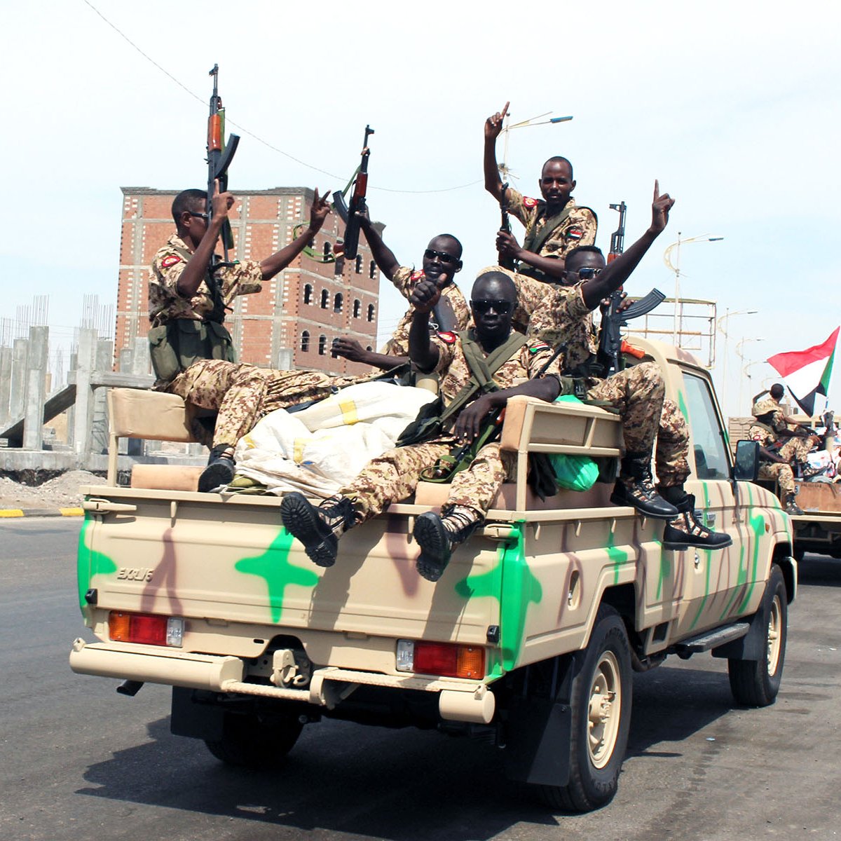 The image depicts a group of armed soldiers riding in the back of a military truck. They appear to be celebrating or expressing excitement, with some of them raising their arms and displaying gestures. The truck has a camouflage pattern, and the background shows a developing urban area, with construction materials and buildings under construction. The overall scene suggests a military convoy or a display of force.