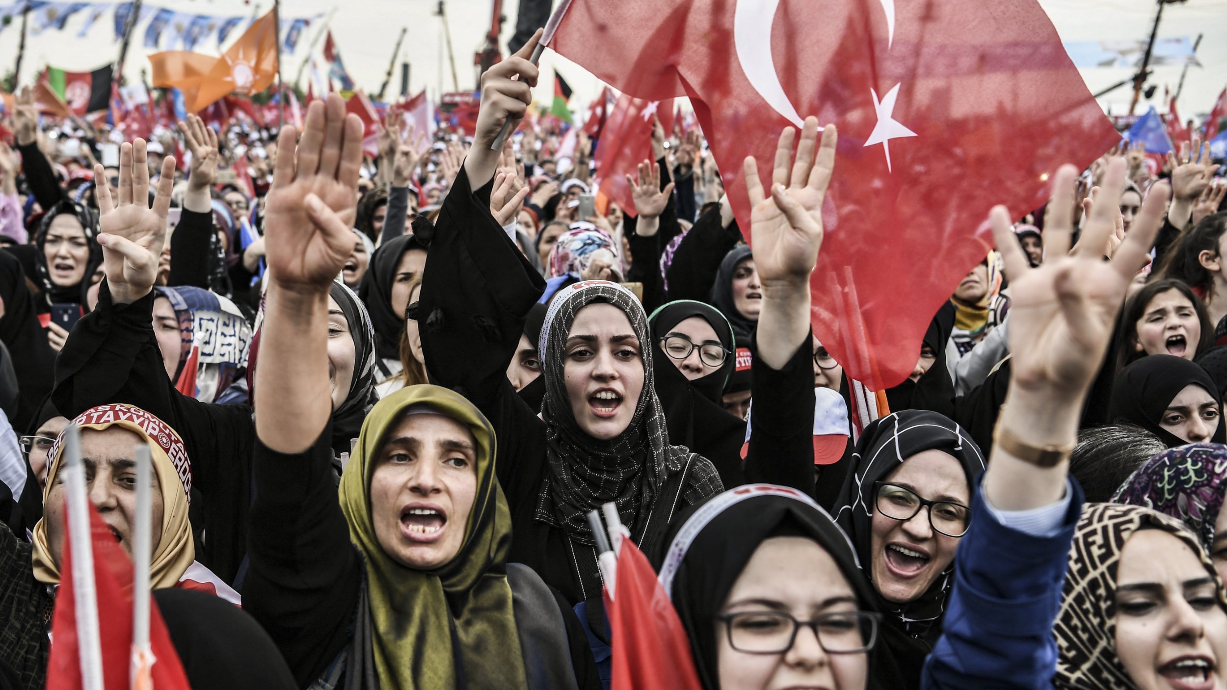 الصورة تظهر حشودًا من النساء في تظاهرة أو فعالية جماهيرية. العديد من النساء يحملن الأعلام التركية ويعبرن عن حماسهن من خلال رفع الأيادي. يبدو أنهن يرتدين ملابس تقليدية أو حجاب، مما يعكس تنوع أساليب التعبير عن الهوية. الجو العام يظهر حيوية وشغفًا، مع وجود لافتات وأعلام في الخلفية تدل على رسالة أو هدف مشترك.