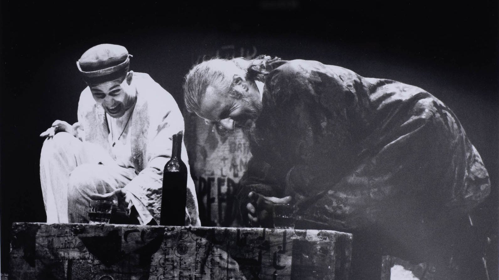 Deux hommes en scène, riant, près d'une bouteille sur une caisse au fond sombre.