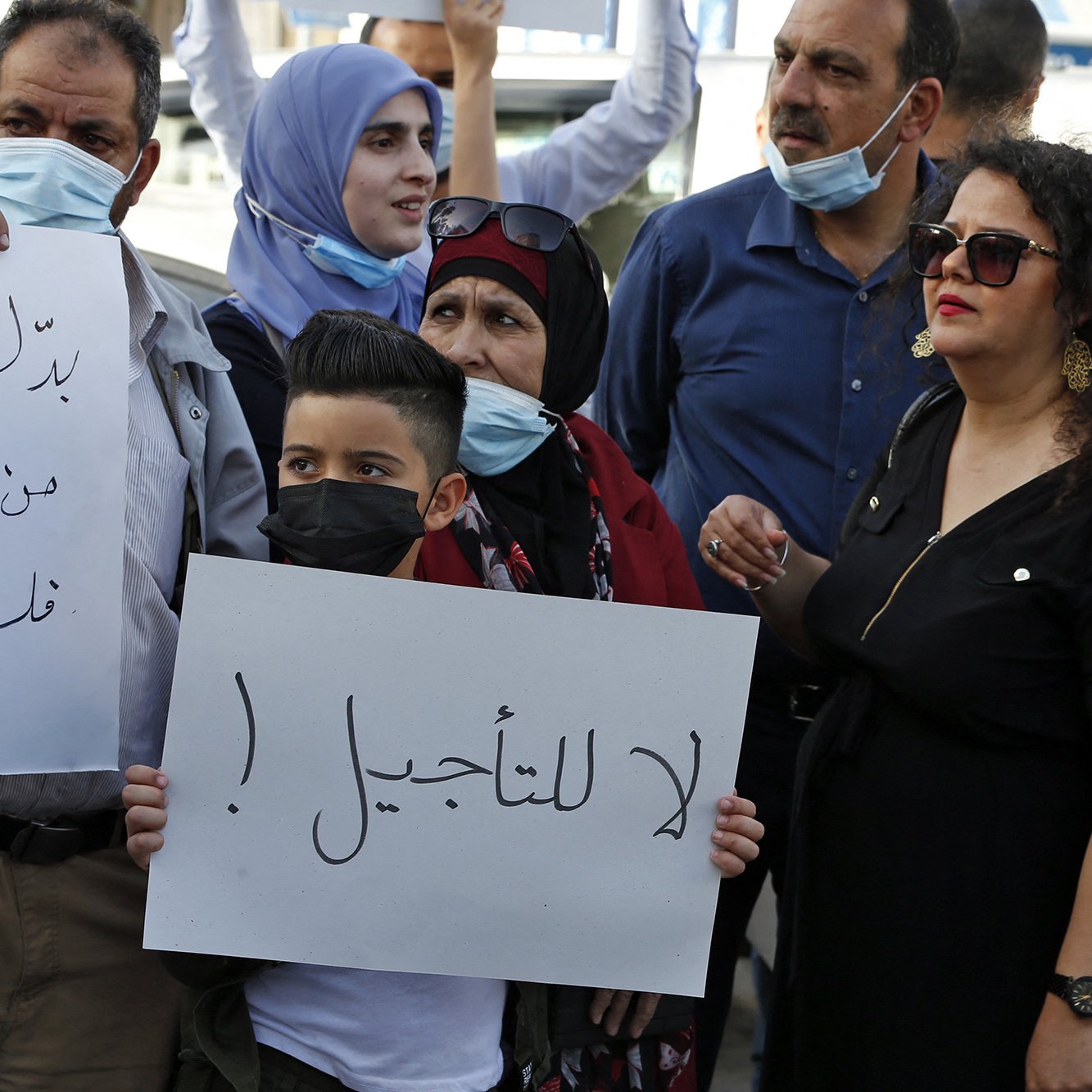در این تصویر، گروهی از افراد در حال تظاهرات هستند. برخی از آنها ماسک به صورت دارند و تابلوهایی در دست دارند. یکی از تابلوها حاوی جمله‌ای است که به زبان عربی نوشته شده و به معنای "نه به تاخیر!" است. افراد در تصاویر شامل زنان و مردانی با پوشش‌های مختلف هستند، و برخی از آنها با چهره‌های جدی و مصمم به بیان خواسته‌های خود پرداخته‌اند. فضای کلی نشان‌دهنده‌ی اعتراض و تلاش برای بیان نظرات و مطالبات است.