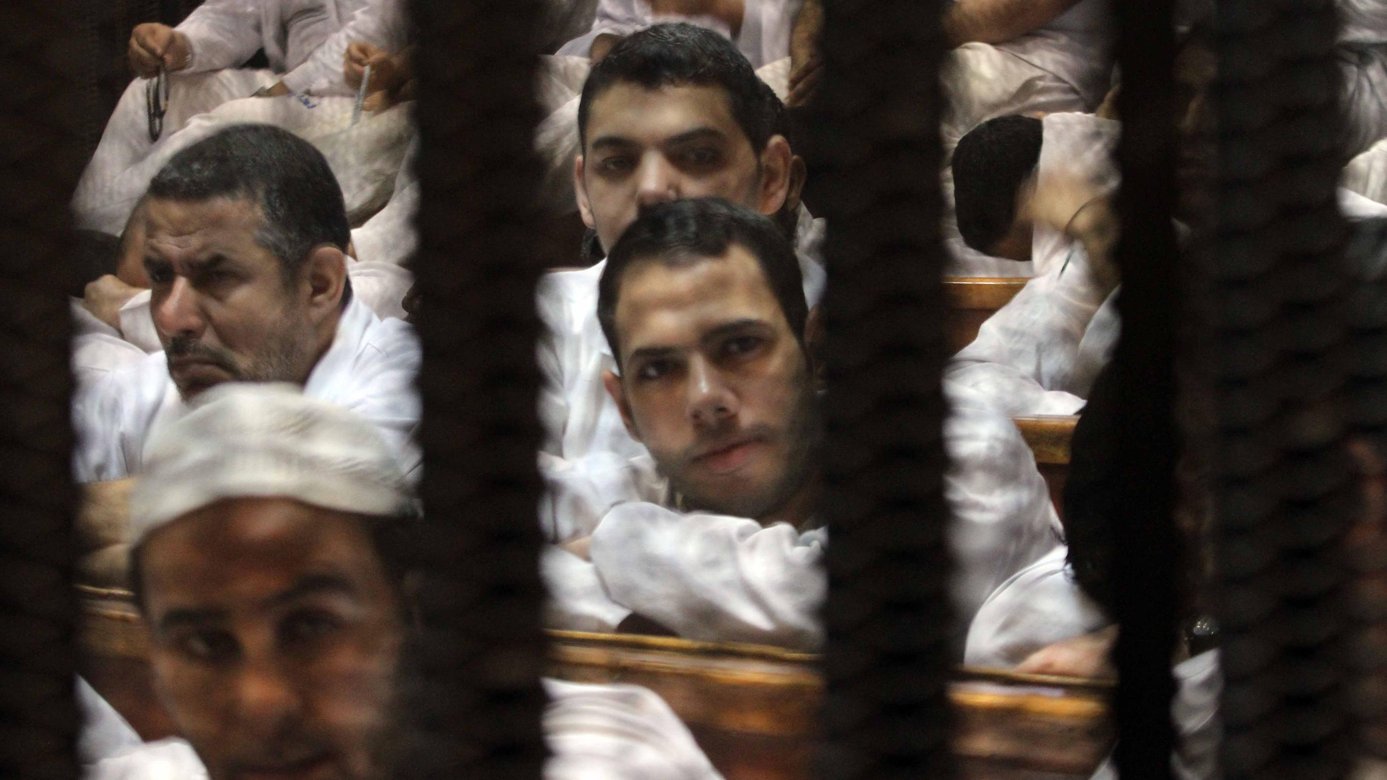 L'image montre un groupe d'hommes assis dans une salle, probablement lors d'une audience judiciaire. Ils portent des vêtements simples, généralement de couleur claire. Leurs expressions semblent sérieuses, et ils sont entourés de barreaux, ce qui suggère un environnement de détention ou de tribunal. L'ambiance générale est tendue, et la composition de la photo cadre bien les visages des hommes à travers les barreaux.