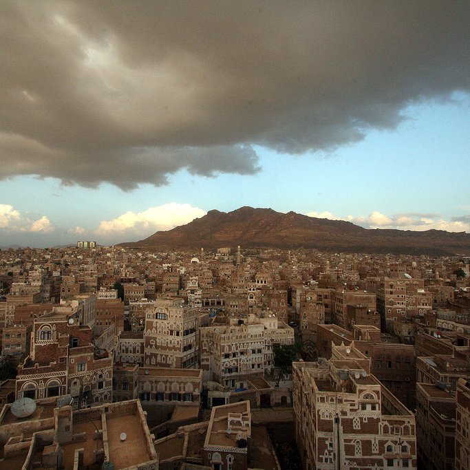 L'image montre une vue panoramique d'une ville traditionnelle, probablement située au Yémen, avec des bâtiments en adobe et des structures architecturales distinctives. On aperçoit des montagnes en arrière-plan, sous un ciel nuageux qui laisse entrevoir quelques éclaircies. La lumière du soleil couchant semble illuminer une partie de la ville, mettant en valeur les détails des constructions. L'ensemble dégage une atmosphère calme et pittoresque, reflétant la beauté du paysage urbain.