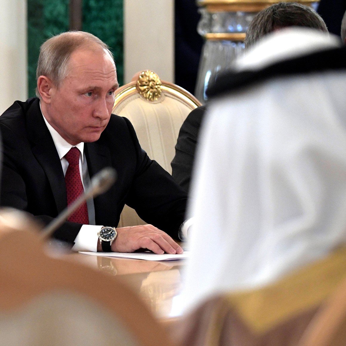 تظهر الصورة اجتماعًا رسميًا حيث يجلس رجل يرتدي بدلة ذات لون داكن. يبدو أنه مركز جداً، حيث ينظر إلى الطاولة أمامه. في الخلفية، يمكن رؤية أشخاص آخرين يرتدون الملابس التقليدية، مما يعطي انطباعًا عن حدث رسمي أو دبلوماسي يشارك فيه ممثلون من ثقافات مختلفة. يحيط بالاجتماع أجواء من الجدية والتركيز.