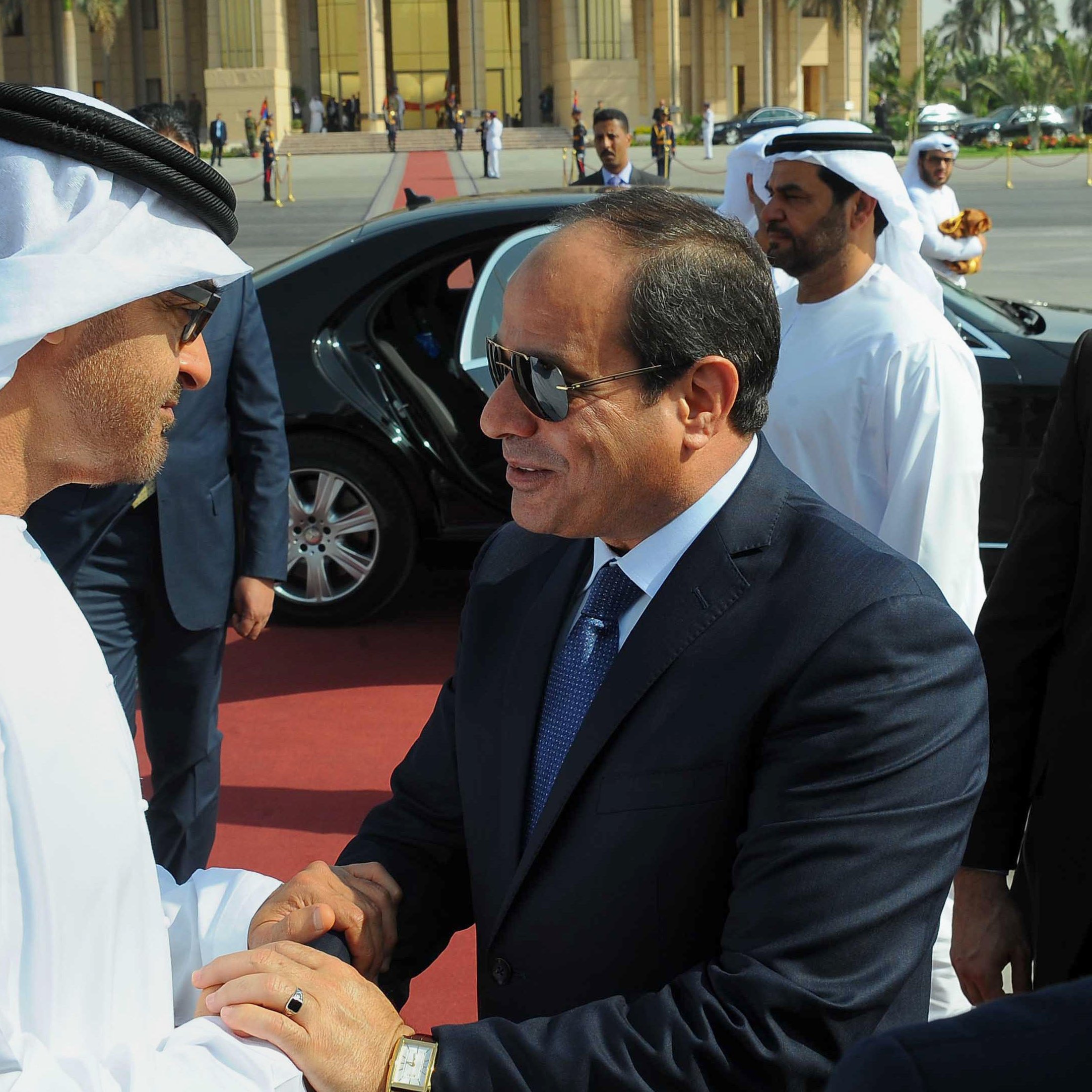 The image depicts two men in formal attire engaged in a handshake, suggesting a cordial greeting. One man is dressed in traditional Emirati clothing, including a white kandura and headscarf, while the other is wearing a dark suit and tie. They appear to be in an official setting, with a luxurious car and a group of well-dressed individuals in the background. The atmosphere indicates a diplomatic or state visit, emphasizing cooperation and respect between the two figures.