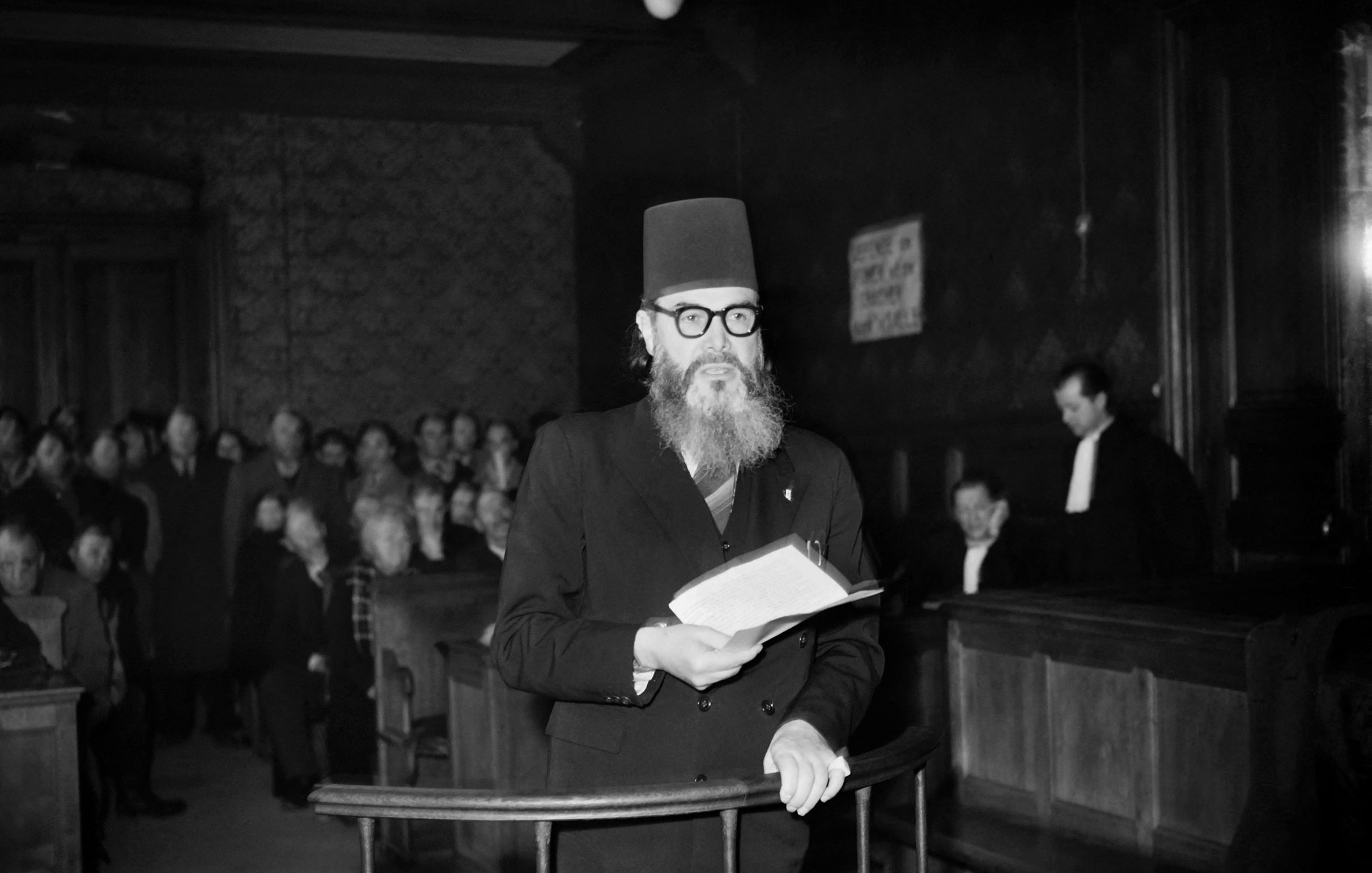 L'image montre un homme portant un turban noir et des lunettes, se tenant debout dans une salle d'audience. Il lit un document, tandis qu'un groupe de personnes l'écoute attentivement en arrière-plan. L'environnement est sombre, avec des boiseries et des bancs de juge visibles. Une personne en toge est également présente, ajoutant à l'atmosphère judiciaire de la scène.