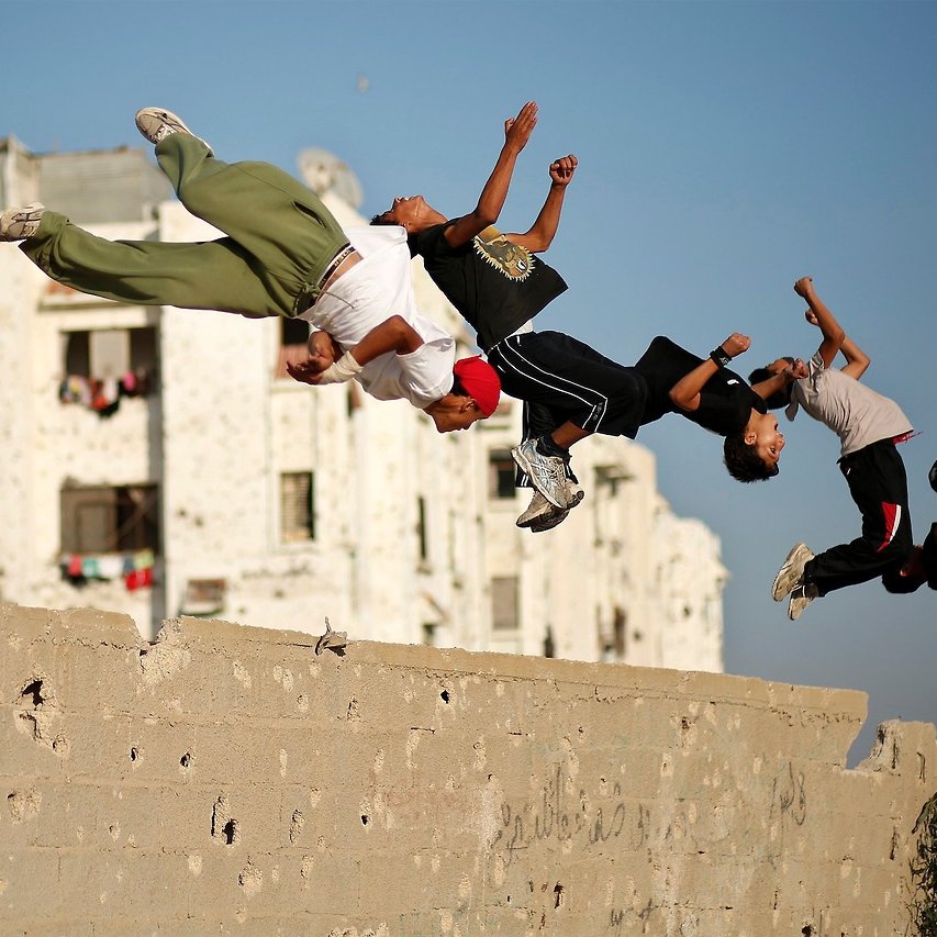 La imagen muestra a un grupo de jóvenes realizando acrobacias o saltos, en un entorno urbano. Están en el aire, ejecutando movimientos como un salto hacia atrás, mientras que de fondo se pueden ver edificios con paredes desgastadas. Los chicos parecen estar disfrutando de la actividad, mostrando energía y alegría. La escena refleja un momento de juego y diversión en un área urbana.