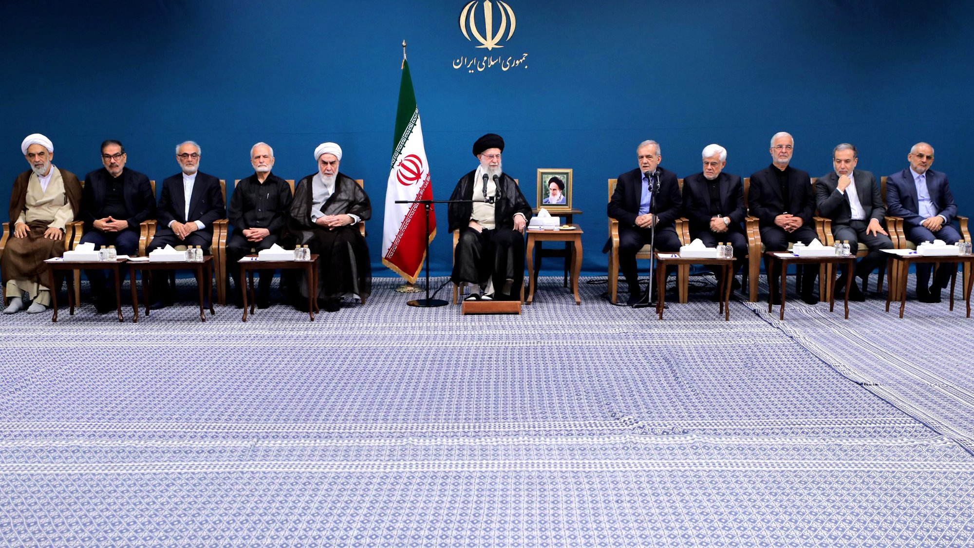 L'image montre un groupe de personnes assises en rangée sur un tapis, dans un environnement formel. En arrière-plan, il y a un drapeau iranien et un portrait d'une figure importante, probablement un leader. Les participants semblent être des hommes en costume sombre et en tenue religieuse, certains portant des turbans. Ils semblent engagés dans une discussion ou une réunion officielle. L'ambiance est solennelle et professionnelle.