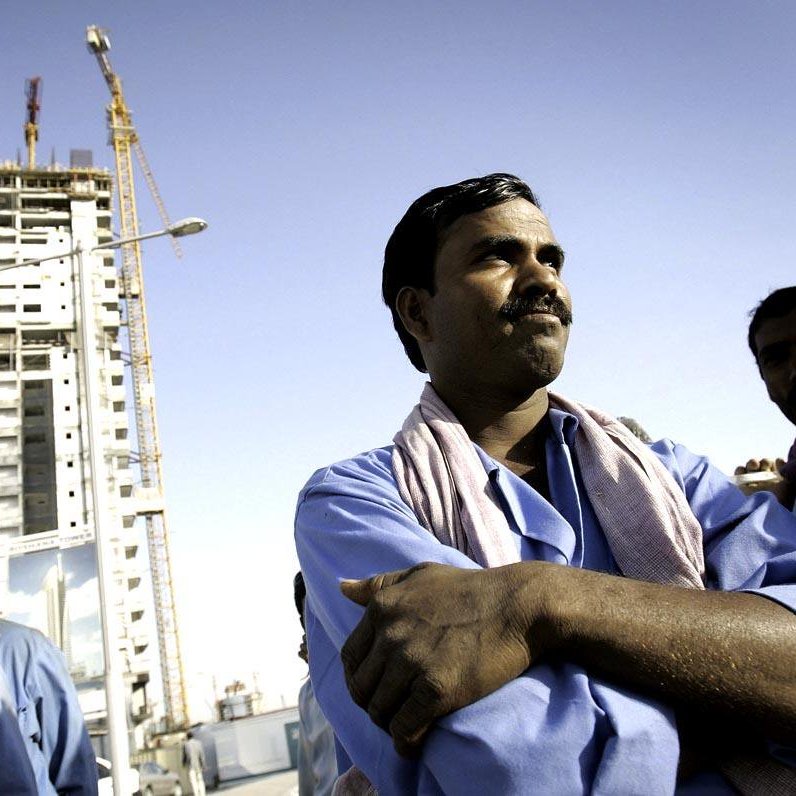 L'image montre un groupe d'ouvriers sur un chantier de construction. Au premier plan, un homme est au centre de l'image, les bras croisés et affichant une expression confiante. À l'arrière-plan, on voit un bâtiment en construction, avec des grues qui travaillent. Les ouvriers portent des vêtements de travail, et certains d'entre eux portent des casques de sécurité. Le ciel est dégagé, suggérant une journée ensoleillée. L'ensemble de la scène évoque l'atmosphère d'un site de construction animé avec des travailleurs engagés dans leur tâches.