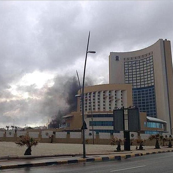 L'image montre un bâtiment moderne de grande taille, probablement un hôtel ou un immeuble de bureaux, situé dans une zone urbaine. À l'arrière-plan, on peut voir de la fumée noire s'élever dans le ciel, ce qui suggère qu'il y a eu un incendie ou une explosion. Le temps semble nuageux, ajoutant une atmosphère sombre à la scène. Des lampadaires bordent la route, et il y a des espaces verts et des plantations autour du bâtiment.