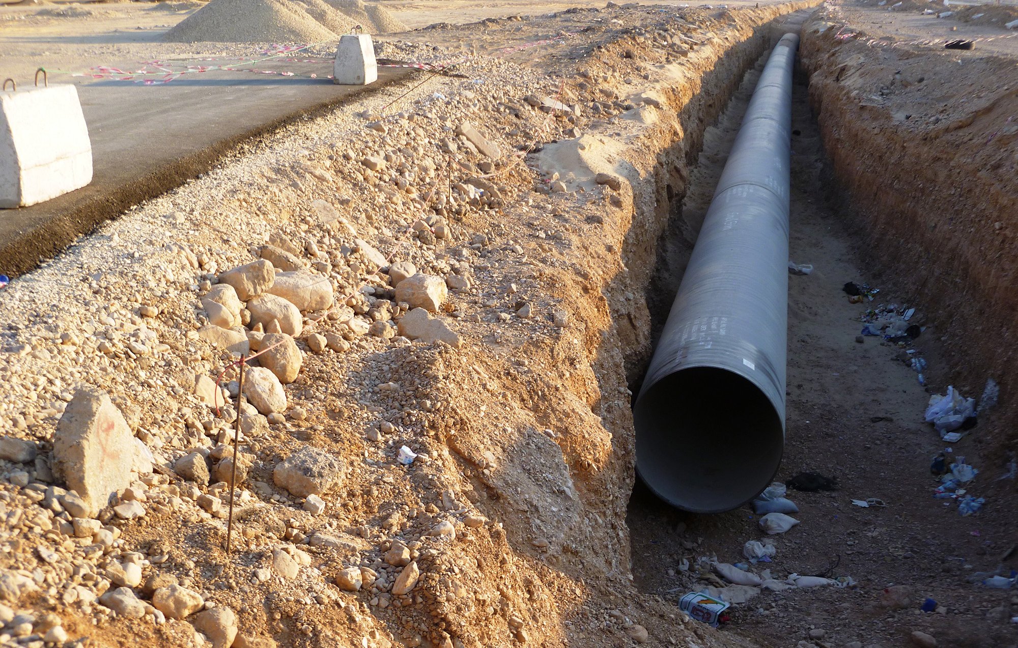 L'image montre un espace de construction où un grand tuyau en métal est disposé dans une tranchée creusée dans le sol. Le terrain environnant est aride et rocailleux, avec quelques débris, comme des bouteilles, dispersés dans la tranchée. Au fond, on peut voir des engins de chantier et une route partiellement construite. L'ambiance générale évoque un projet d'infrastructure en cours.