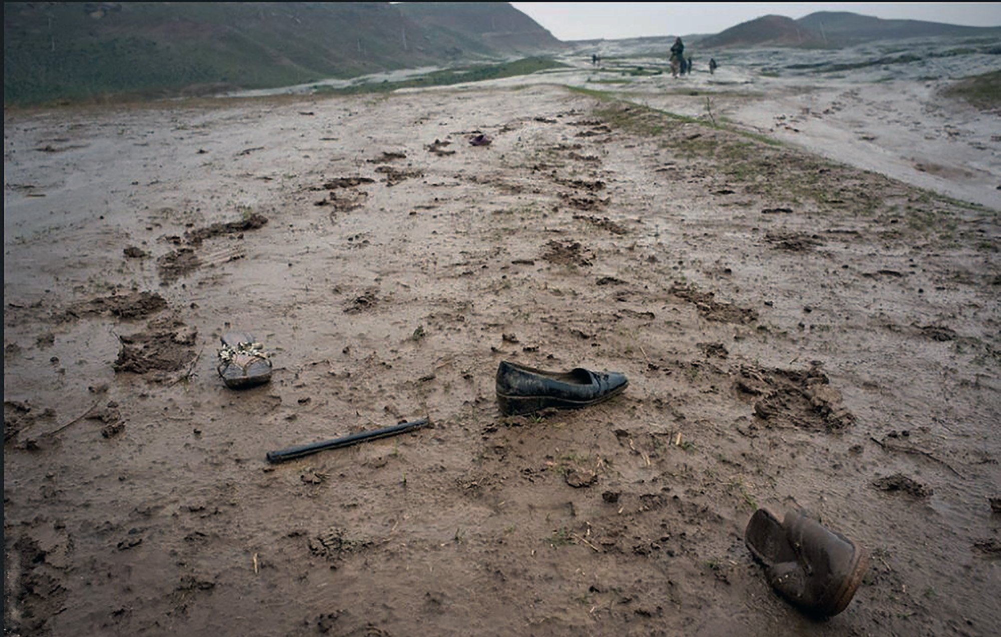 L'immagine mostra un paesaggio desolato, con un terreno fangoso e bagnato. Ci sono alcune scarpe abbandonate sulla strada, di cui una sembra essere un tacco alto e l'altra una scarpa più semplice. Sul suolo si notano segni di fango e detriti, creando un'atmosfera di degrado e abbandono. Sullo sfondo, si possono vedere delle figure lontane che camminano, suggerendo un contesto di difficoltà o di transito in un ambiente difficile. La scena comunica un senso di solitudine e trascuratezza.