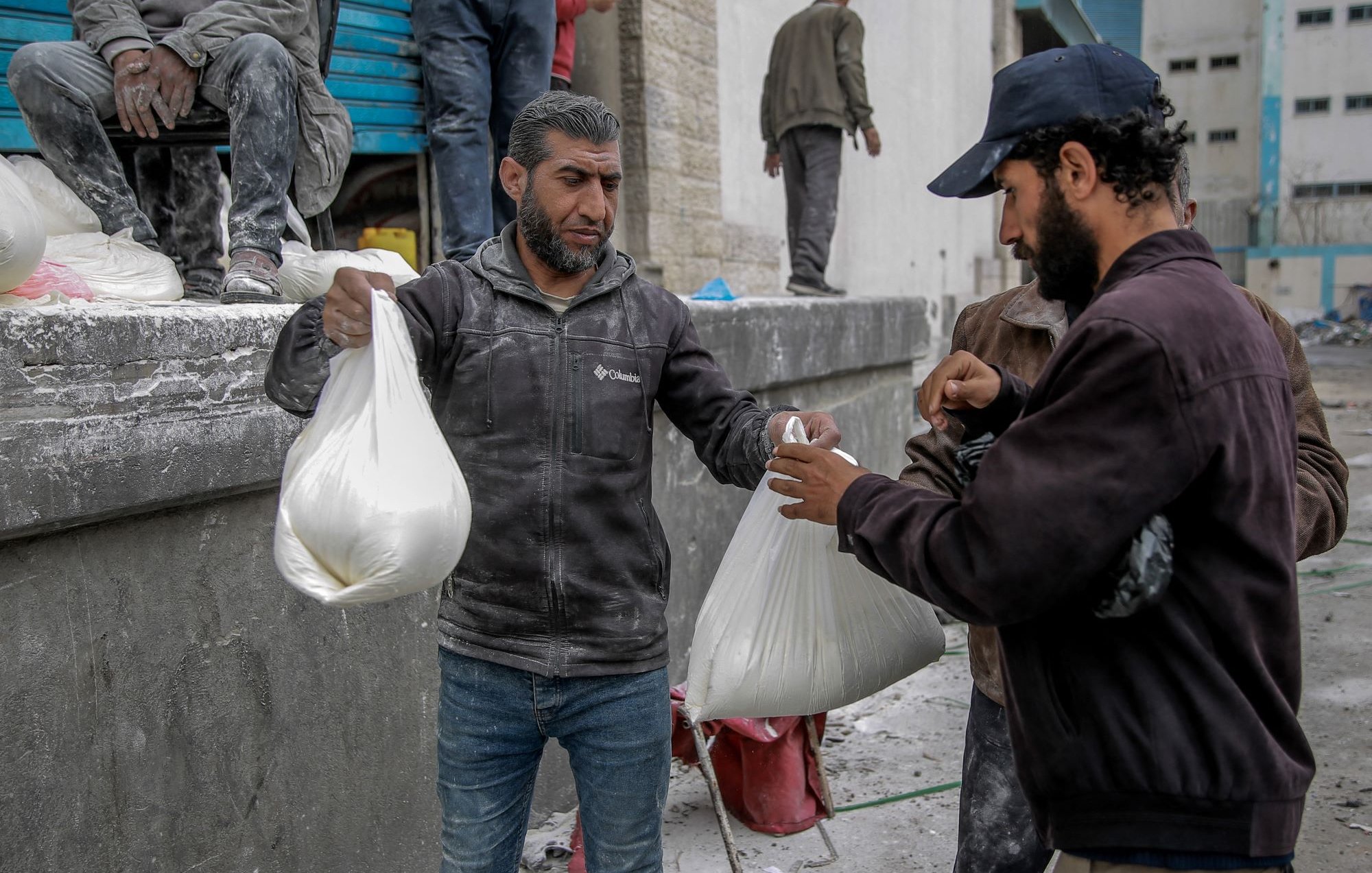 L'image montre des hommes dans un environnement urbain, apparemment en train de distribuer ou de recevoir des sacs. Ils semblent porter des vêtements de travail, certains étant couverts de poussière ou de matériau. L'ambiance suggère une activité liée à l'aide humanitaire ou à une distribution de biens, dans un contexte peut-être de reconstruction ou d'assistance. Les interactions entre les hommes semblent concentrées et sérieuses.