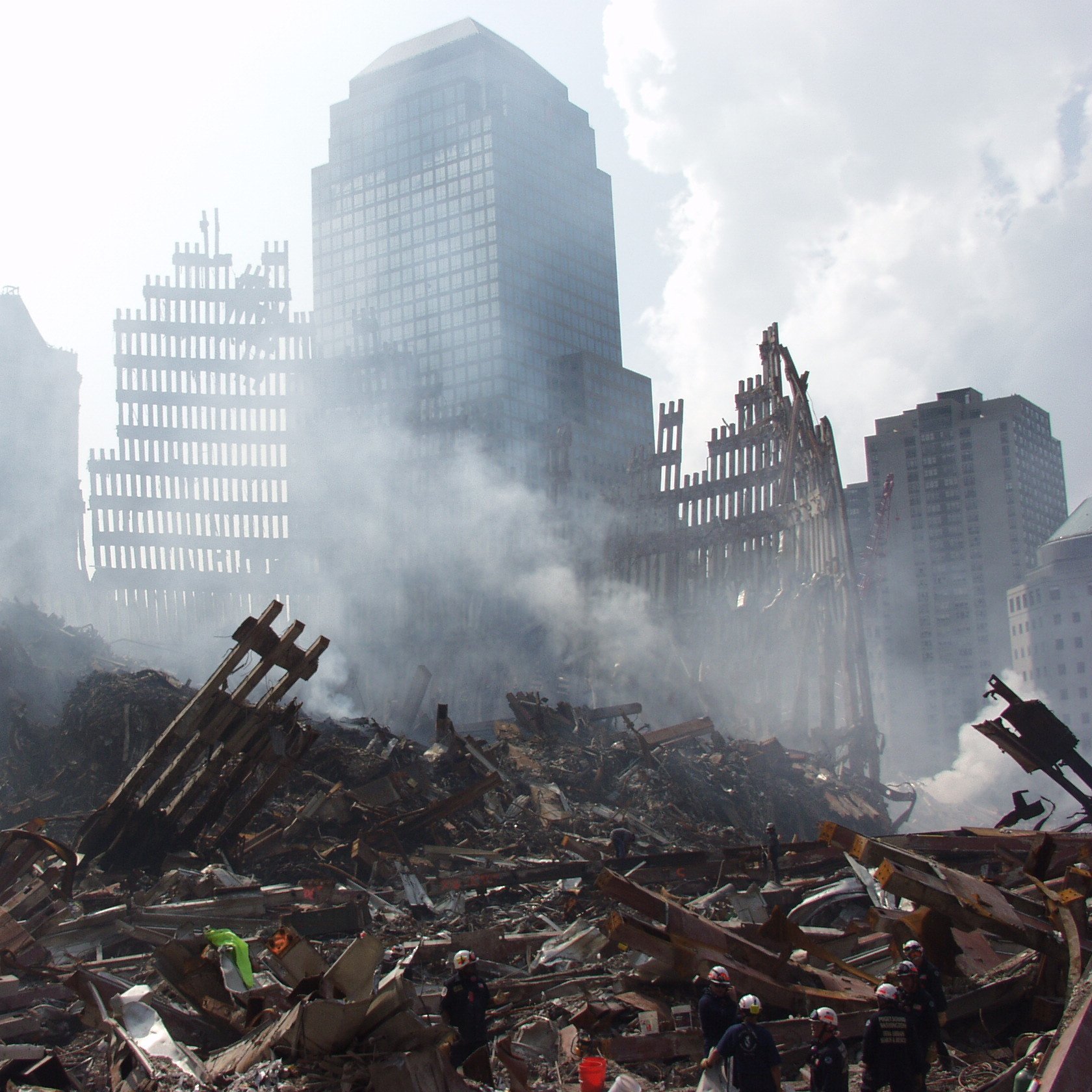 تصویر مربوط به یک صحنه خرابی و ویرانی است. در پس‌زمینه، ساختمان‌های بلند و ناتمام دیده می‌شوند که برخی از آنها به شدت آسیب‌دیده‌اند. در جلوی تصویر، خرابه‌هایی از سازه‌ها و تیرک‌های فلزی با دودی که از میان آنها بلند می‌شود، مشاهده می‌شود. حالت کلی تصویر نشان‌دهنده یک حادثه ویرانگر است و فضای ملتهب و غم‌انگیزی را ایجاد می‌کند.