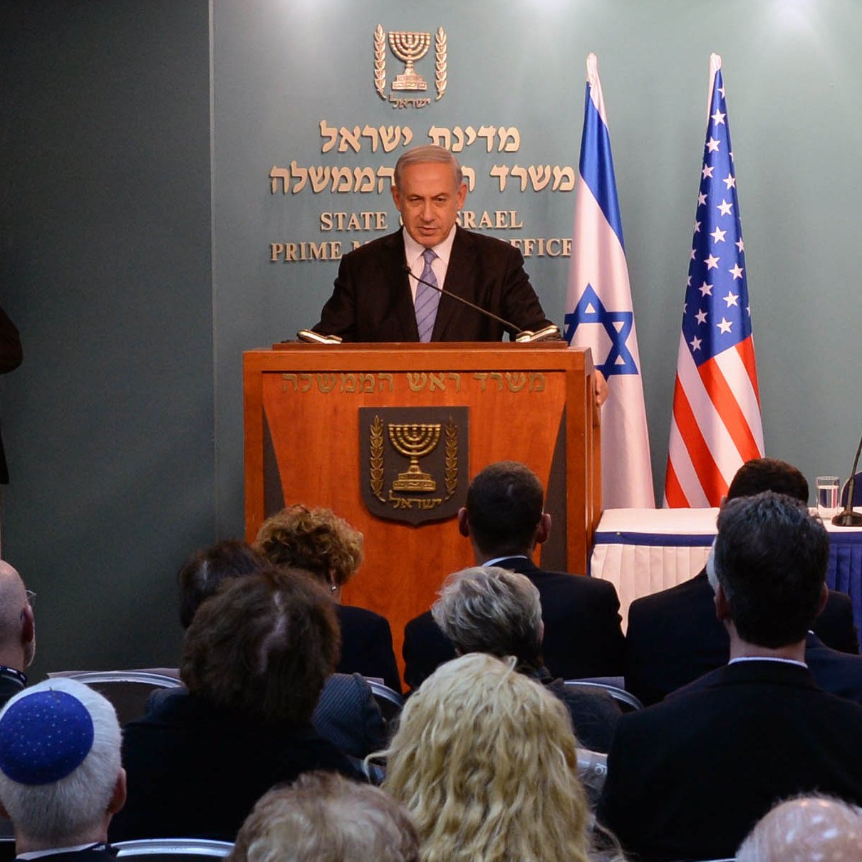 L'image montre un homme en train de s'adresser à un public depuis un podium marqué du sceau d'Israël et des États-Unis. Il y a des personnes assises derrière lui, écoutant son discours. On peut voir des drapeaux israélien et américain en arrière-plan. L'ambiance semble formelle, probablement dans le cadre d'une conférence ou d'une réunion officielle.