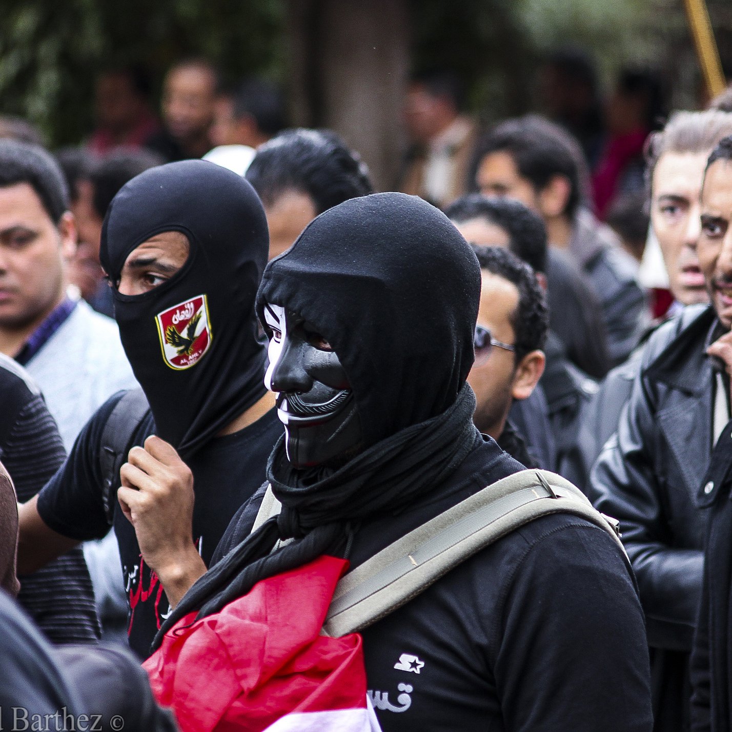 L'image montre un groupe de personnes rassemblées, probablement lors d'une manifestation. Certains portent des masques noirs, tandis que d'autres affichent des expressions déterminées. On peut observer un mélange de vêtements allant des vestes en cuir à des écharpes. L'ambiance semble être à la fois sérieuse et engagée, avec des signes de solidarité entre les participants.