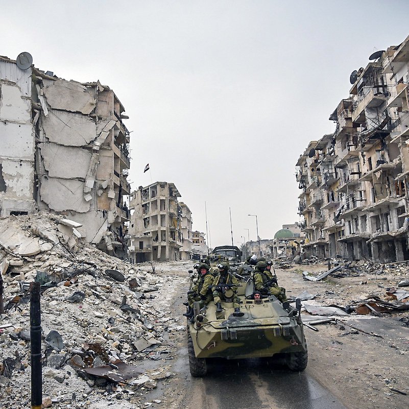 L'image montre une scène de destruction urbaine, probablement à la suite d'un conflit. On peut voir des bâtiments en ruine, avec des façades effondrées et des débris éparpillés sur le sol. Au premier plan, il y a un véhicule militaire, sur lequel des soldats sont présents. L'atmosphère semble sombre et chargée, soulignant les ravages causés par la guerre.