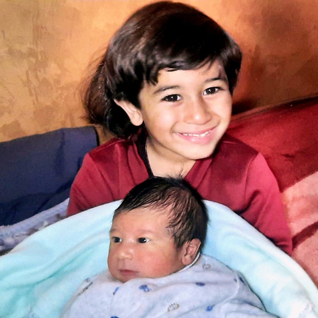 تصویر یک پسر بچه‌ی حدود ۵ ساله را نشان می‌دهد که لبخند می‌زند و در آغوش او یک نوزاد خوابیده است. پسر بزرگ‌تر لباس قرمز پوشیده و نوزاد در یک پتوی آبی پیچیده شده است. پس‌زمینه تصویر رنگی گرم دارد.