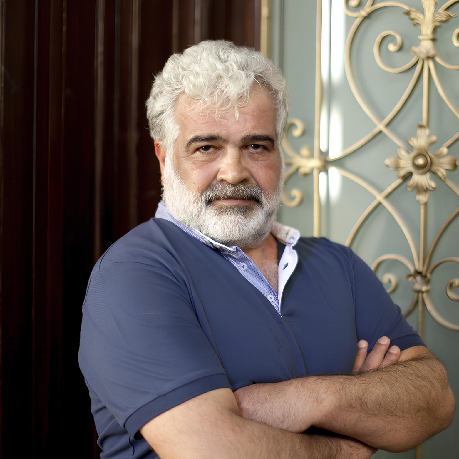 In questa immagine, si vede un uomo maturo con capelli bianchi e una barba folta. Indossa una maglietta blu e ha un'espressione seria, con le braccia incrociate. Sullo sfondo, ci sono elementi decorativi di metallo che aggiungono un tocco elegante all'ambiente. La luce mette in risalto i dettagli del suo viso e dell'abbigliamento.