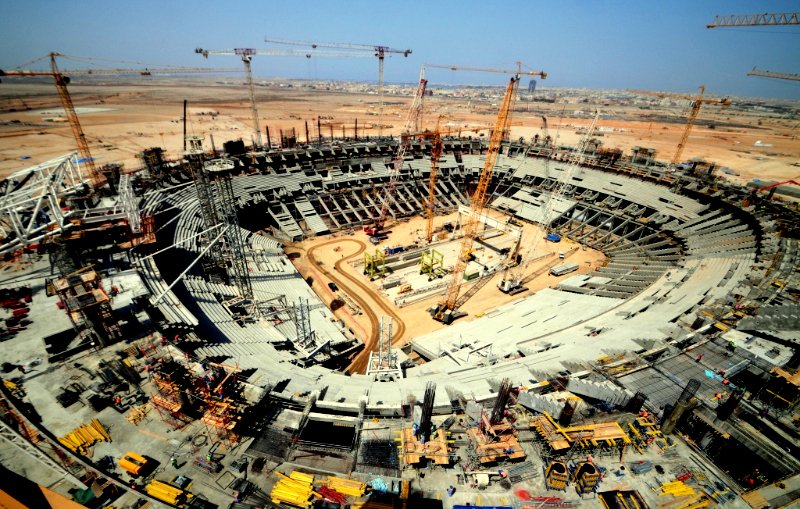 L'image montre un immense chantier de construction d'un stade. On peut voir une grande arène en forme de cercle, avec des gradins en cours d'assemblage. De nombreuses grues sont présentes, surélevant des matériaux de construction, et divers engins lourds sont dispersés sur le site. À l'horizon, on aperçoit des bâtiments et un ciel dégagé, indiquant une propreté et un bon déroulement des travaux. L'ensemble de la scène illustre l'activité dynamique et l'envergure d'un projet architectural ambitieux.