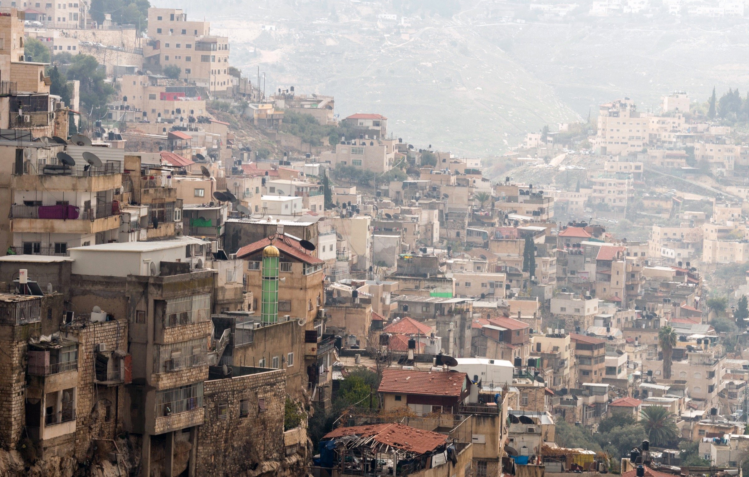 The image shows a densely populated hillside area with a mix of residential buildings. The structures vary in height and architectural style, with many appearing stacked closely together. Some roofs are visible, displaying a range of materials, from tiles to concrete. In the background, more houses and buildings can be seen on the sloping terrain, suggesting a continuation of urban development. The overall scene has a somewhat hazy atmosphere, possibly due to distance or weather conditions.