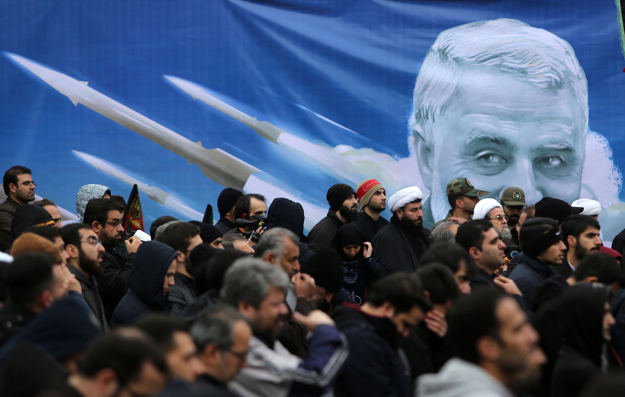 تصویر نشان‌دهنده تجمعی از افراد در یک مراسم است. مردم بیشتر لباس‌های تیره پوشیده‌اند و برخی از آن‌ها در حال گوش دادن به سخنان یا تماشای یک برنامه هستند. در پس‌زمینه، تصویری بزرگ از چهره‌ای که شبیه یک شخصیت شناخته‌شده است وجود دارد و در اطراف آن تصاویری از موشک‌ها دیده می‌شود. این تصویر به نوعی نمادین به نظر می‌رسد و می‌تواند به مناسبت‌های ملی یا مذهبی مربوط باشد. 