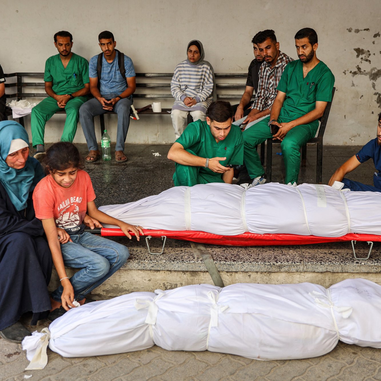 در این تصویر، تعدادی از افراد در یک مکان عمومی نشسته‌اند. در جلو، دو بدن پوشیده شده در پارچه‌های سفیدی قرار دارند. افراد حاضر در تصویر با ظاهری نگران و غمگین به نظر می‌رسند. برخی از آنها لباس‌های پزشکی به تن دارند و به وضوح تحت تأثیر شرایط عاطفی و وضعیت حاضر قرار گرفته‌اند. فضا به طور کلی حاکی از یک وضعیت بحرانی و دردناک است.