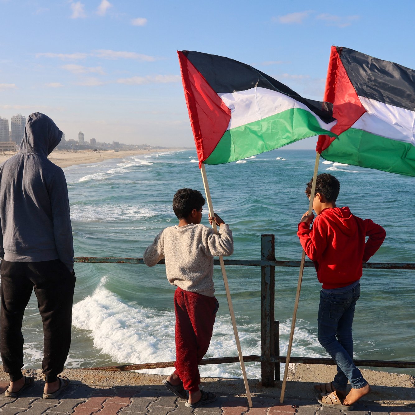 Nell'immagine, tre ragazzi sono in piedi su un molo che si affaccia su una spiaggia e sul mare. Due di loro tengono in alto delle bandiere palestinesi, che sono di colore nero, bianco, verde e rosso. Il ragazzo più piccolo, in una felpa grigia, sembra osservare attentamente l'orizzonte. La scena è illuminata da una luce naturale, con il cielo blu e alcune nuvole sparse. Le onde del mare si infrangono dolcemente sulla riva, creando un'atmosfera tranquilla. Gli altri due ragazzi indossano abbigliamento casual e sembrano condividere un momento di riflessione insieme.