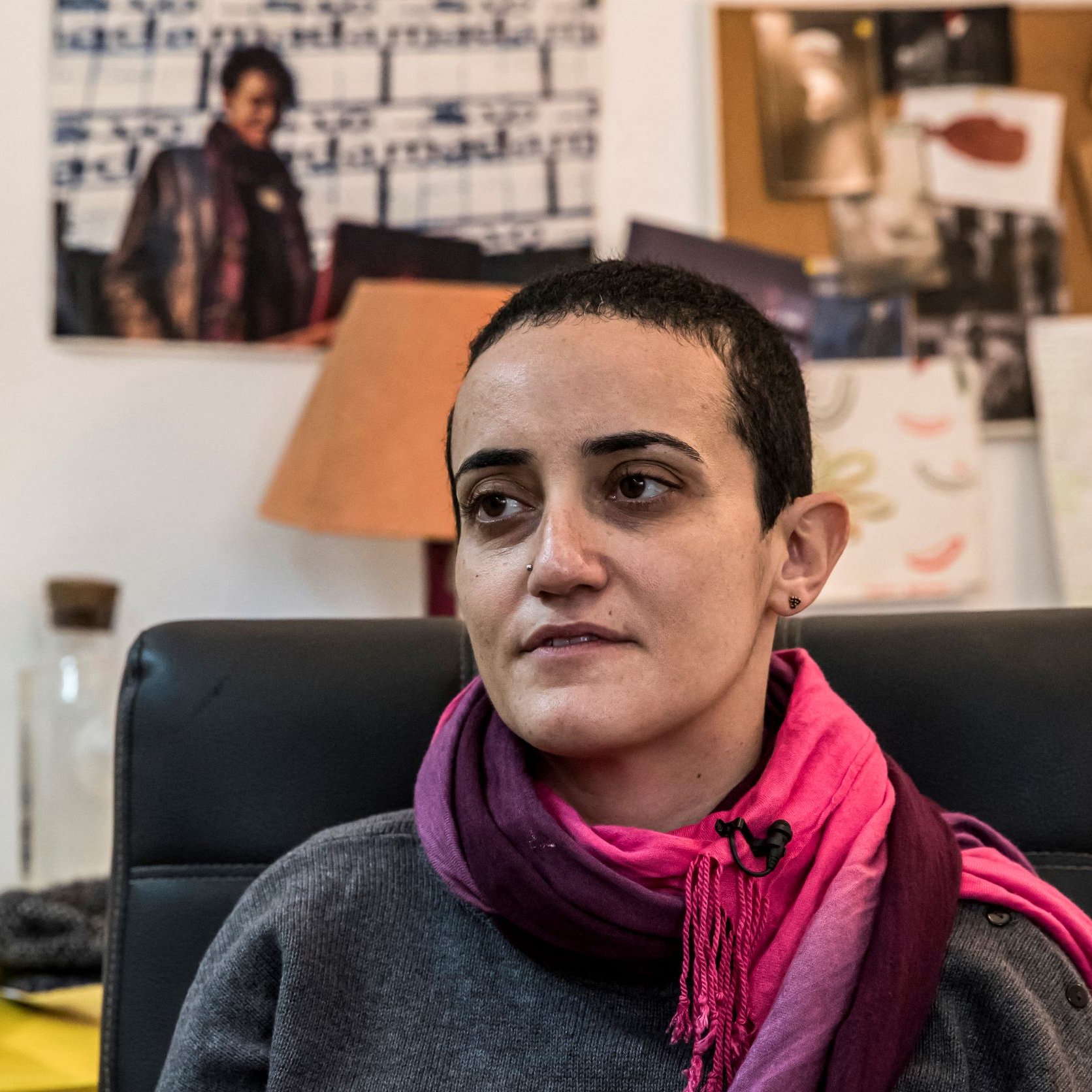 Je ne sais pas qui est présent sur cette image. Cependant, je peux te dire que la personne semble assise dans un bureau, portant un pull gris et une écharpe rose. On peut voir un environnement de travail avec des documents et des photos affichées en arrière-plan. L'éclairage donne une ambiance chaleureuse à la scène.