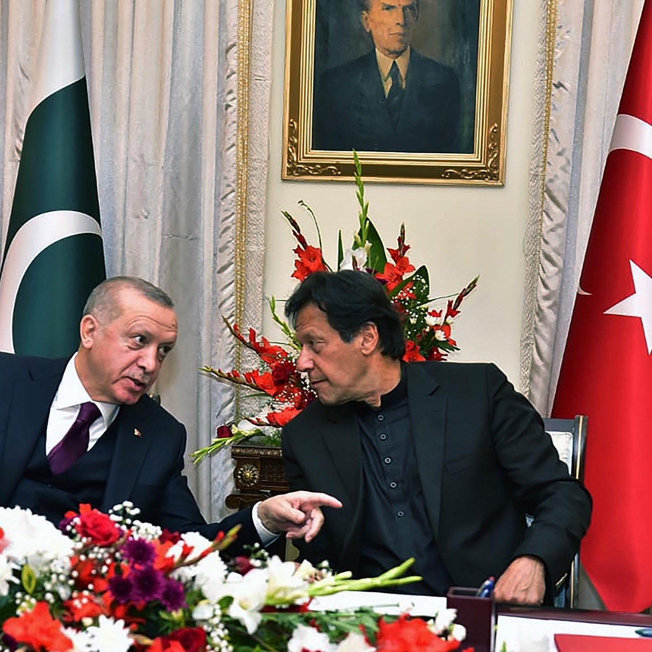 En la imagen se pueden ver a dos hombres conversando en un ambiente formal, rodeados de banderas de Turquía y Pakistán. Hay un arreglo floral colorido en la mesa, y detrás de ellos se encuentra un retrato en la pared. La escena transmite un ambiente de diplomacia y colaboración entre los dos países.