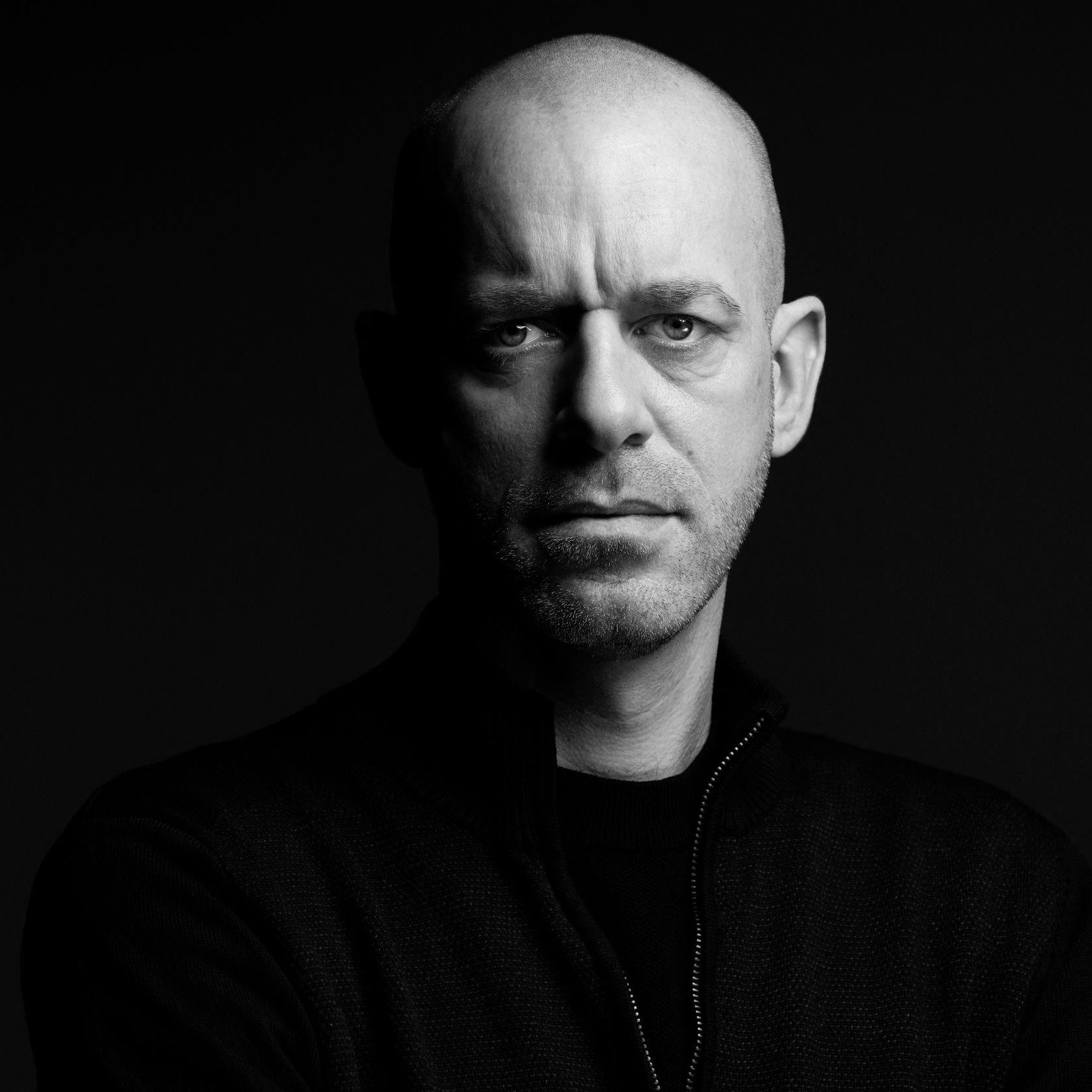 الصورة تُظهر رجلًا ذو رأس أصلع، يقف في ضوء خافت مما يبرز ملامح وجهه بوضوح. عيونه تحمل تعبيرًا جادًا وقويًا، بينما يحدق بشكل مباشر نحو الكاميرا. يرتدي سترة داكنة، ويبدو أن الخلفية سوداء، مما يضيف إلى جمالية الصورة ويعطي إحساسًا بالدراماتيكية.