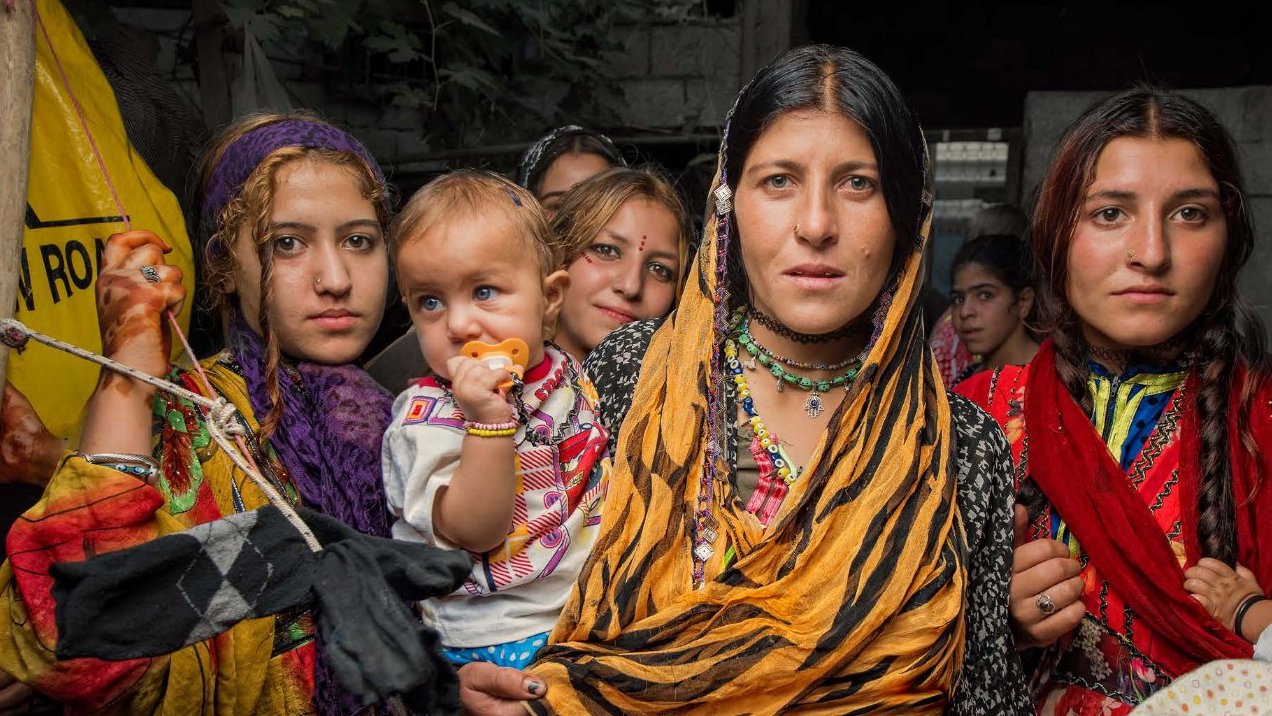L'image montre un groupe de femmes et d'enfants, probablement dans un environnement rural. Les femmes portent des vêtements colorés et traditionnels, avec des foulards sur la tête. L'une d'elles tient un bébé dans ses bras. Les expressions sur leurs visages sont variées, allant de la curiosité à la détermination. On peut aussi voir des éléments culturels dans leur tenue, renforçant l'idée d'une riche tradition. L'arrière-plan semble un peu flou, suggérant un cadre rural ou communautaire.