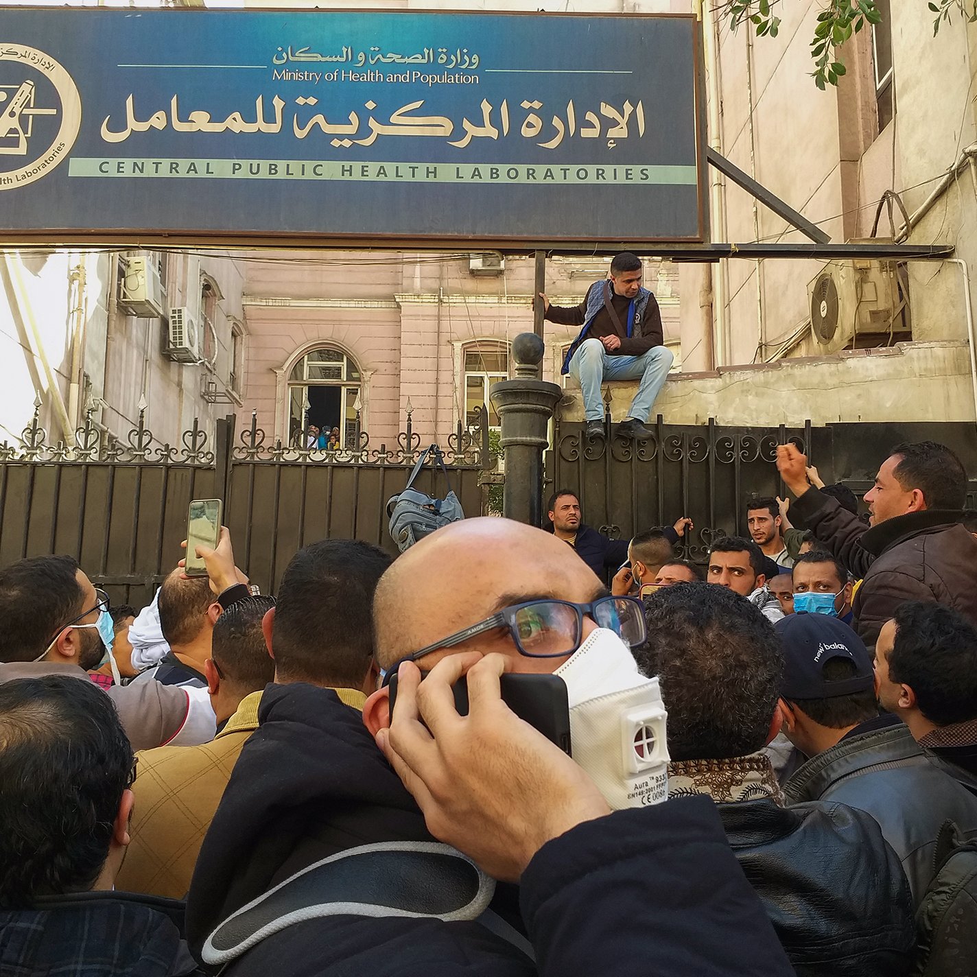 L'image montre une foule rassemblée devant un bâtiment marqué par une enseigne en arabe et en anglais, indiquant qu'il s'agit des laboratoires de santé publique. Des hommes sont visibles, certains portant des masques, tandis qu'un individu est assis sur une barrière. L'atmosphère semble tendue ou agitée, et les personnes semblent discuter ou crier. Le cadre urbain suggère un contexte de mobilisation ou de manifestation.