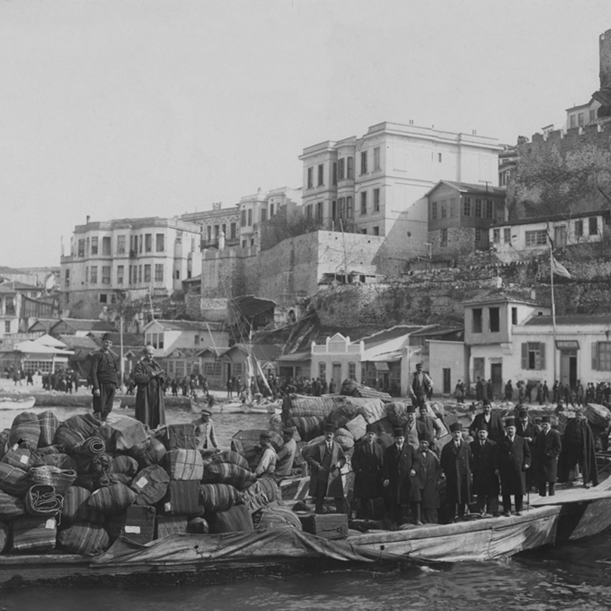 La imagen muestra un puerto animado en un contexto histórico, con un barco de madera cargado de bultos y barriles en primer plano. Un grupo de personas, posiblemente trabajadores o comerciantes, está de pie sobre la embarcación. Al fondo, se puede ver una serie de edificios y una muralla de piedra que se eleva, sugiriendo la presencia de una ciudad costera. La escena tiene un aspecto antiguo, lo que sugiere que fue tomada en una época pasada, posiblemente a finales del siglo XIX o principios del XX. Las actividades portuarias y el diseño arquitectónico transmiten una sensación de vida cotidiana en el puerto.
