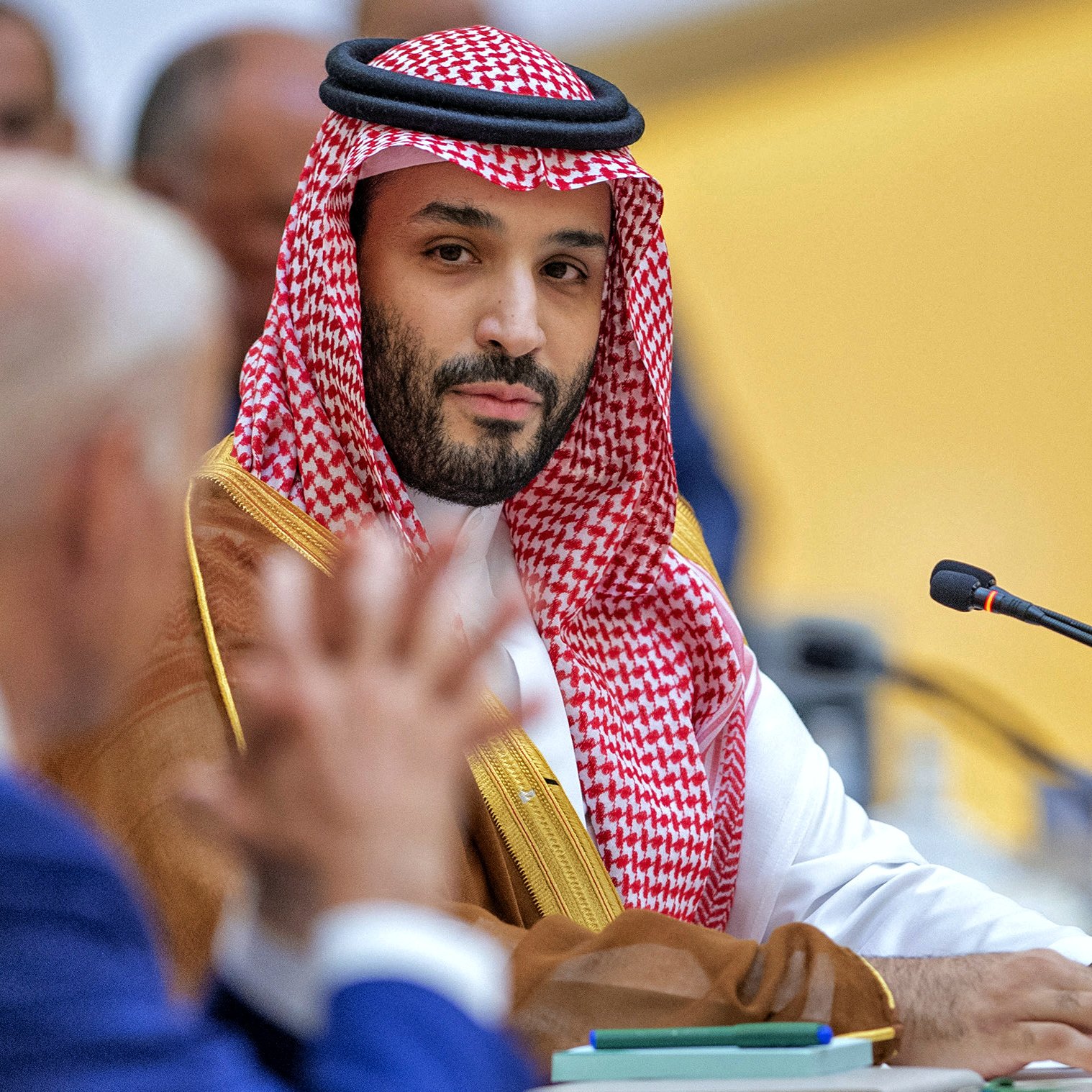 این تصویر مربوط به یک نشست یا جلسه رسمی است. در سمت چپ، یک مرد با لباس رسمی و کت و شلوار آبی نشسته است که به نظر می‌رسد در حال صحبت کردن است. در سمت راست، یک مرد دیگر با لباس سنتی عربی و عمامه قرمز و سفید، در حال گوش دادن به سخنان فرد مقابل است. پس‌زمینه سالن جلسه به طرزی طراحی شده که نشان‌دهنده یک محیط رسمی و دیپلماتیک است.