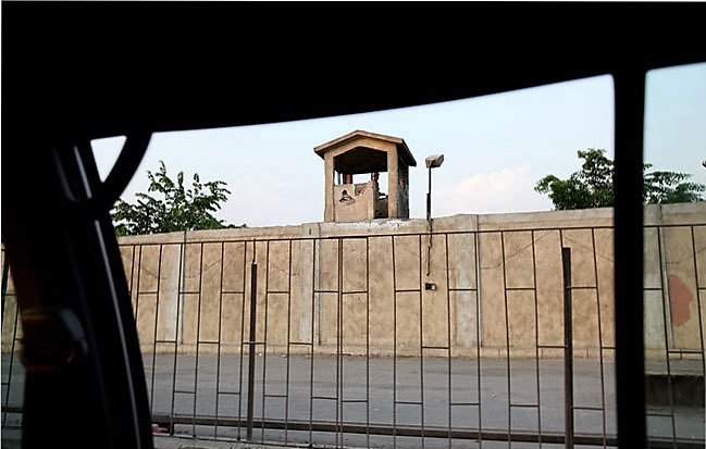 تظهر في الصورة جزء من سياج عازل يحيط بمكان ما، مع وجود برج مراقبة صغير في الأعلى. يمثل البرج نقطة مراقبة، بينما الأسفل يظهر حاجزًا يفصل بين الشارع والفضاء المحيط. تضيء السماء بلونٍ فاتح، مما يدل على أن الوقت قد يكون في فترة المساء أو الصباح. يبدو أن الصورة قد تم التقاطها من داخل مركبة، حيث يظهر إطار السيارة من الجهة اليسرى.