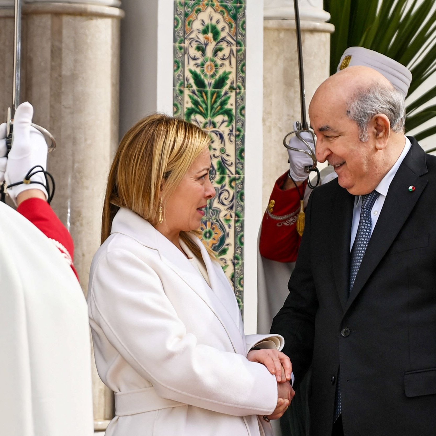 Nell'immagine si possono vedere due persone che si stringono la mano in un contesto formale. Una delle persone indossa un cappotto bianco e ha un sorriso amichevole, mentre l'altra è vestita in modo elegante, con un atteggiamento accogliente. Sullo sfondo si intravedono dettagli architettonici e persone in abiti tradizionali, creando un'atmosfera cerimoniale. La scena riflette un incontro ufficiale, suggerendo un momento di cordialità e dialogo.