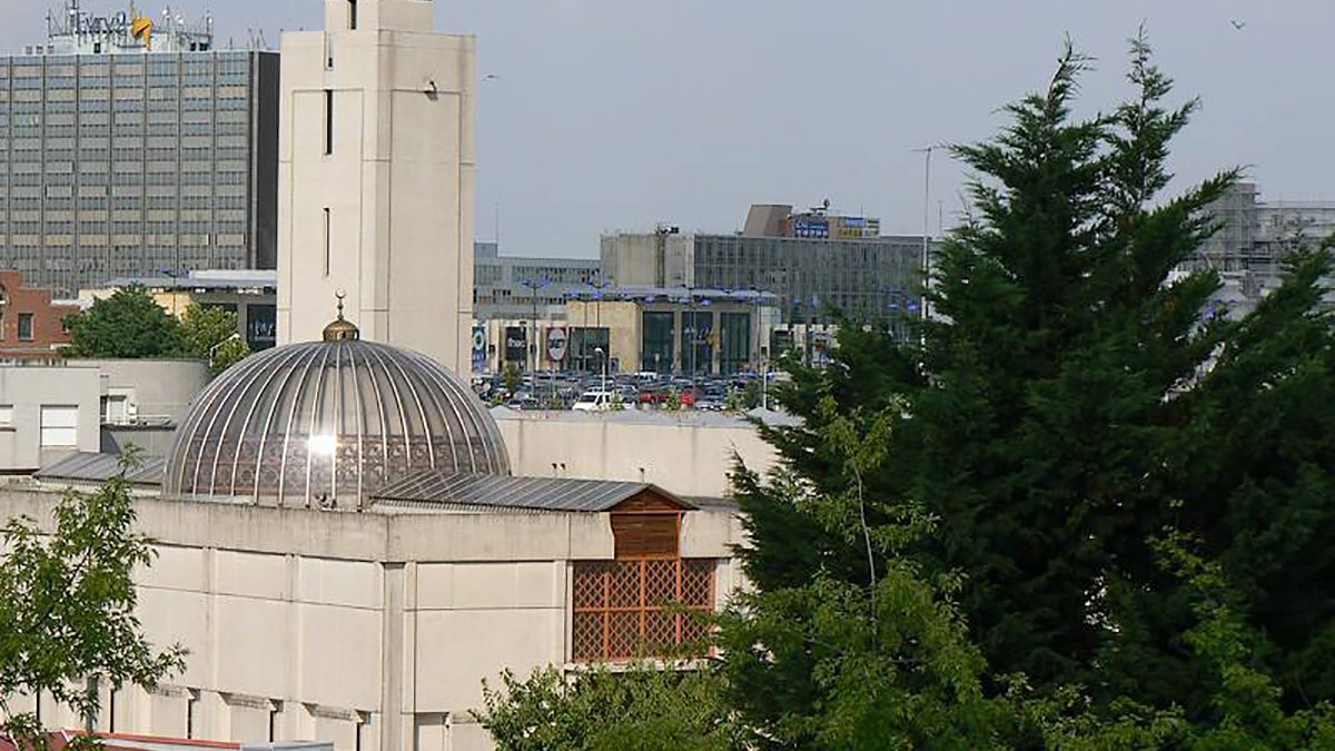 L'image montre un bâtiment architectural avec un dôme et une tour horloge, entouré d'arbres et d'autres structures urbaines en arrière-plan. On peut voir un environnement mixte, combinant des éléments d'architecture moderne et traditionnelle, avec des stations de transports et des bâtiments de bureaux. La scène est bien éclairée, suggérant une journée ensoleillée.