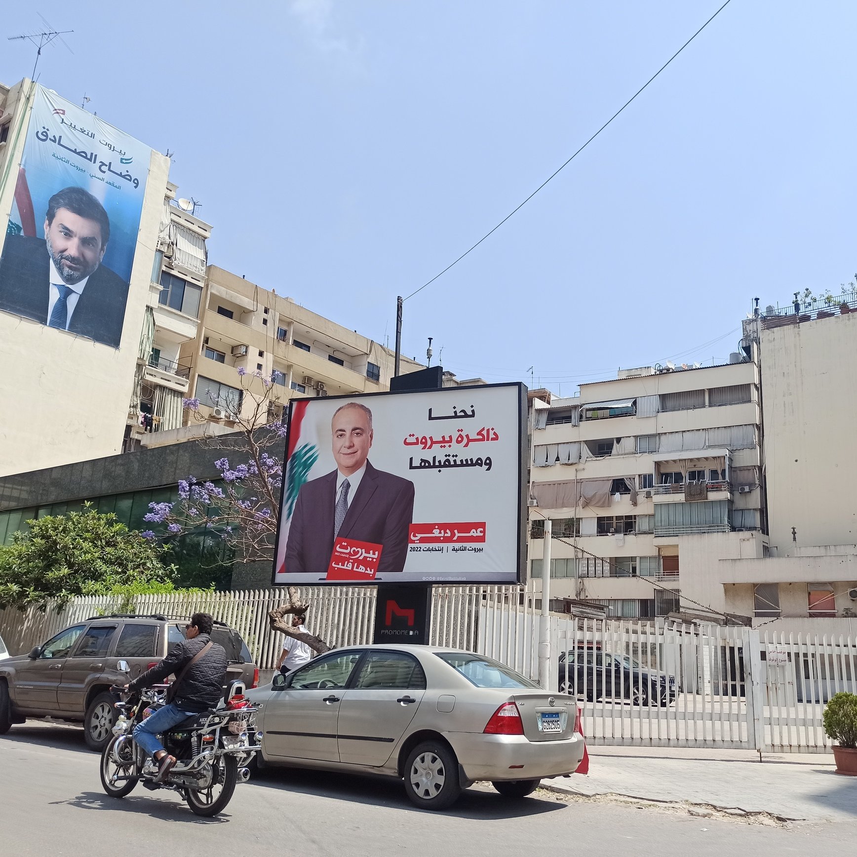 L'immagine mostra una scena urbana con un edificio signorile e un grande cartellone pubblicitario. Il cartellone presenta un ritratto di un uomo in giacca e cravatta, accompagnato da un testo in arabo. Accanto, si può notare un altro edificio che ospita un altro cartellone, ma parte del suo contenuto non è leggibile. Ci sono anche alcune automobili parcheggiate e un motociclista che passa. La via sembra essere molto luminosa grazie al sole.