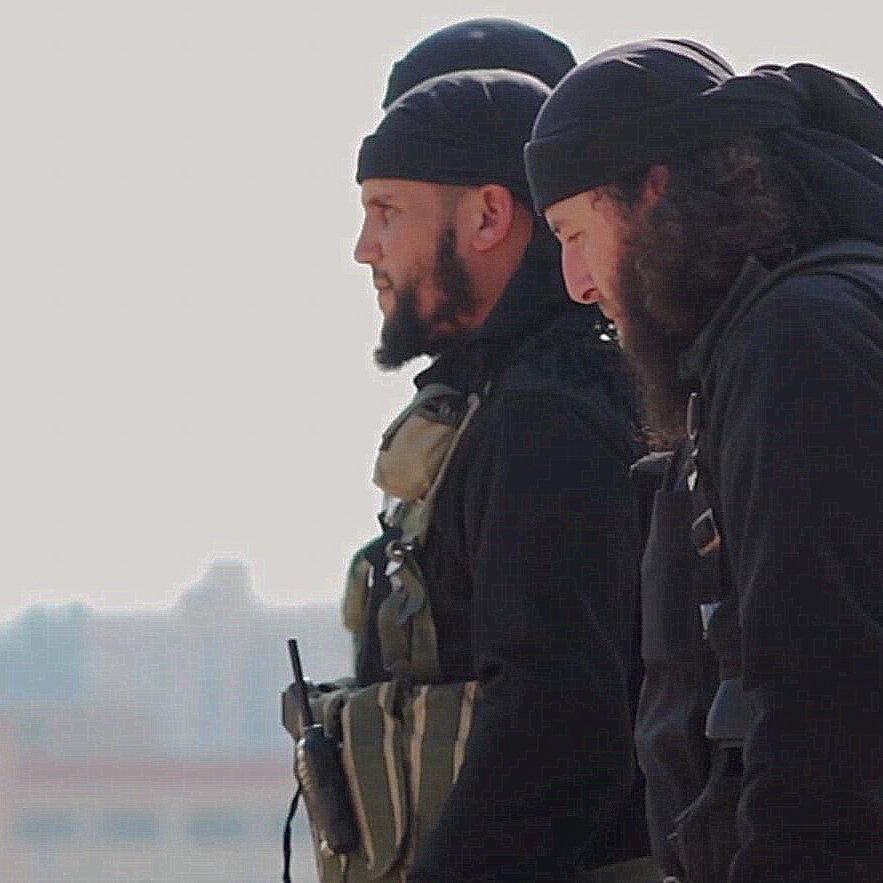 تظهر الصورة مجموعة من الرجال يرتدون ملابس داكنة. يبدو أنهم في وضعية استعداد، حيث يمكن رؤية بعضهم من الجانب. يرتدي البعض منهم شارات أو معدات تكتيكية، مما يدل على أنهم قد يكونون في سياق عمل يتعلق بالأمن أو العمليات العسكرية. خلفهم يظهر مشهد غير واضح، لكن يمكن رؤية بعض المباني في الخلفية. الأجواء عامة تبدو جادة.