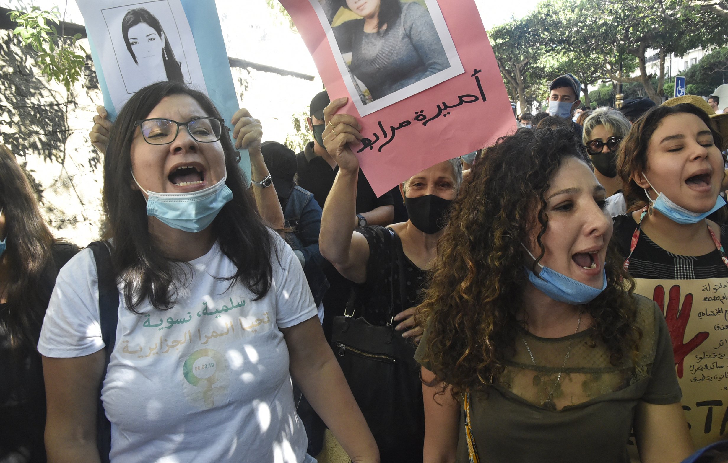 The image depicts a group of women participating in a protest. They are holding up posters, some of which feature photographs of individuals. The women appear to be chanting or shouting, expressing their emotions passionately. Many of them are wearing face masks, likely due to health precautions. The background suggests an outdoor setting with trees, indicating that the demonstration is taking place in a public space. The atmosphere seems charged with activism and determination.
