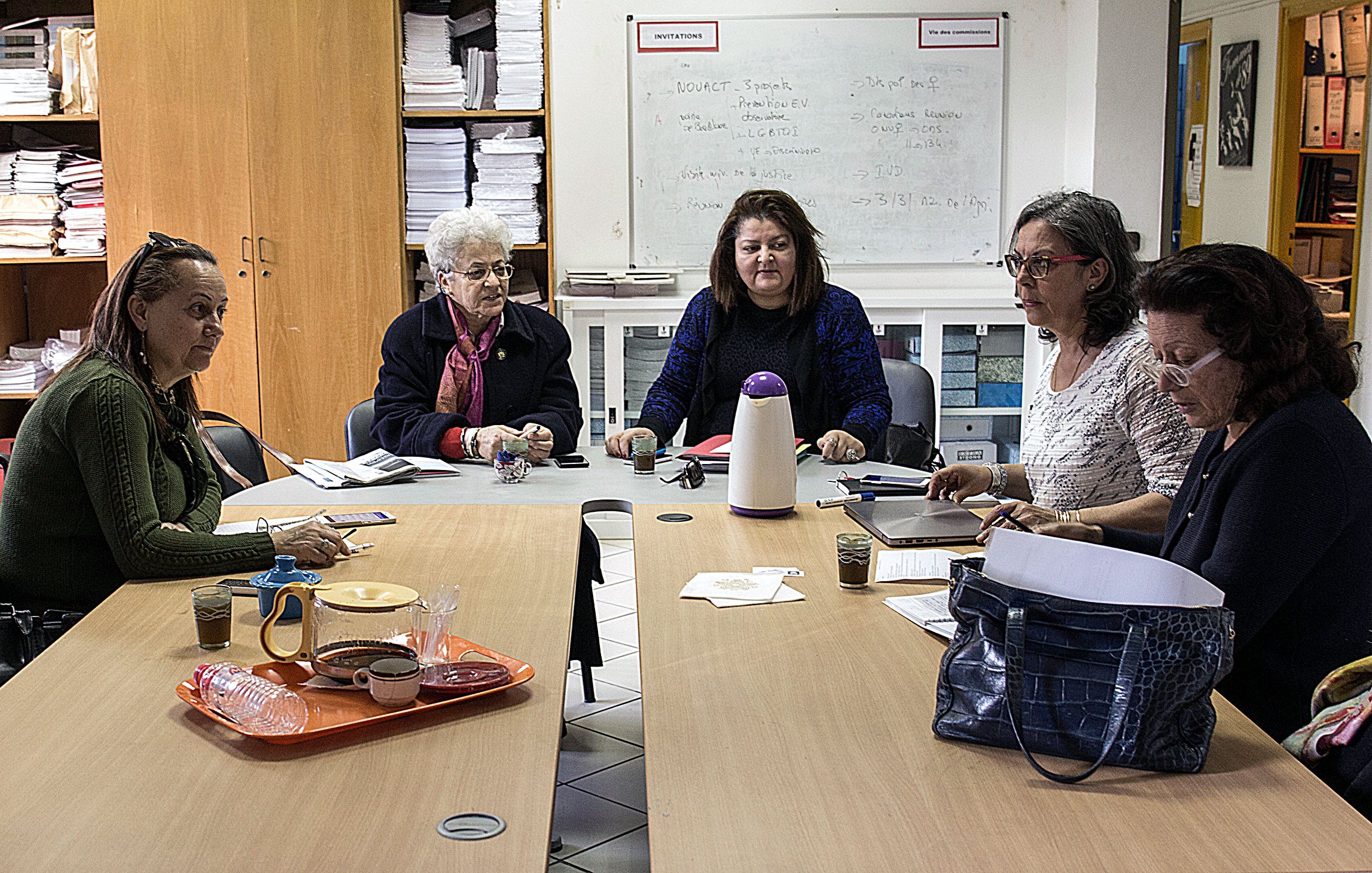 في الصورة، نرى مجموعة من النساء يجلسن حول طاولة في غرفة تحتوي على رفوف مملوءة بالأوراق والمستندات. يظهر على الطاولة بعض الأكواب والأطعمة الخفيفة، بينما يبدو أن المشاركات في اجتماع أو جلسة مناقشة. النساء يبدين مهتمات بما يحدث، حيثيزن التفاعل مع بعضهن البعض. الغرفة تملك أجواء مهنية، مع جدران بيضاء وسبورة خلفهن.