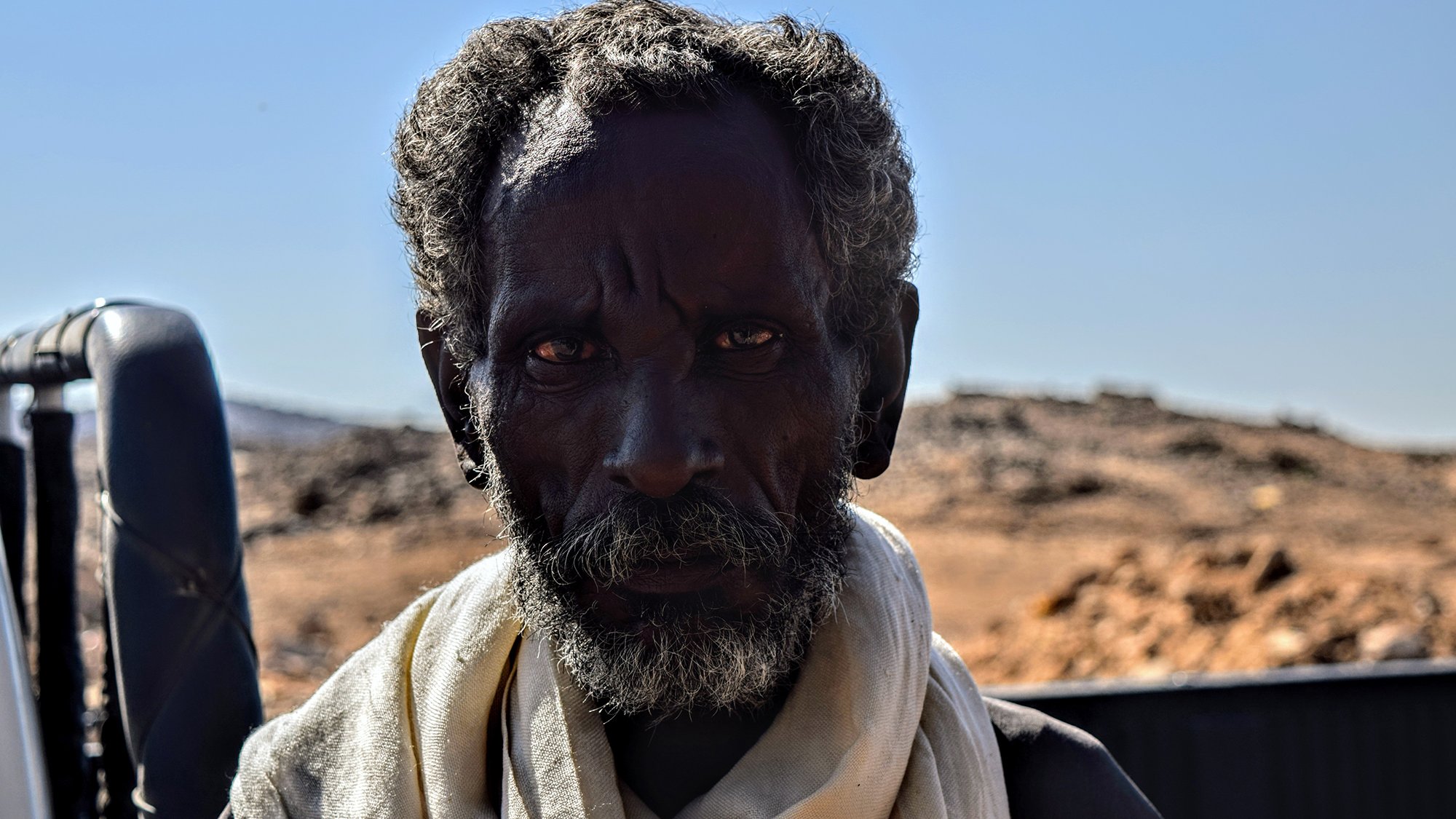L'image montre un homme avec une peau foncée et des cheveux crépus, portant une écharpe claire autour du cou. Son regard est intense et semble exprimer une certaine sagesse ou expérience. L'arrière-plan est flou et semble représenter un paysage désertique avec des montagnes ou des collines à distance. La lumière est vive, ce qui met en valeur les traits du visage de l'homme.