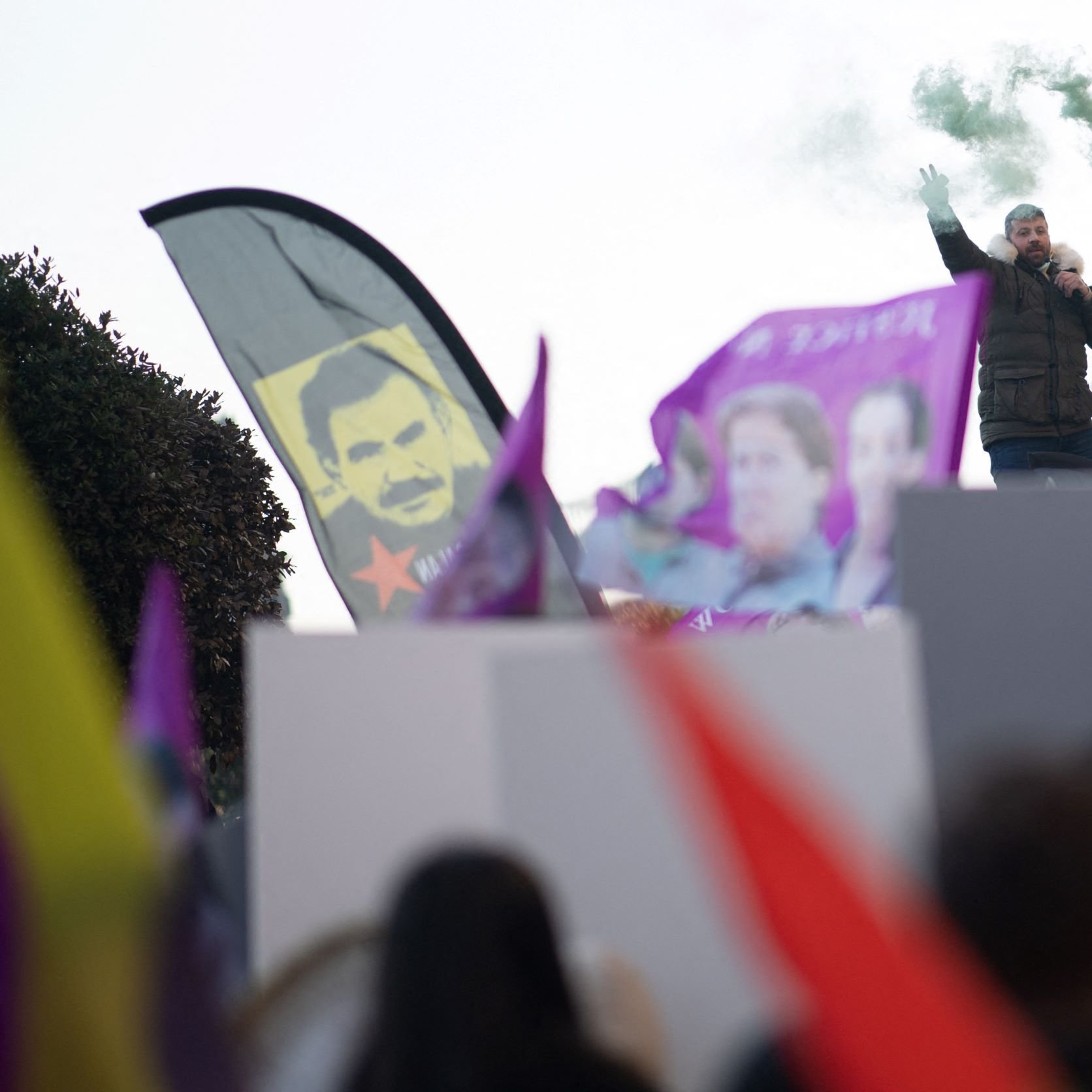 در این تصویر، تعدادی از افراد در حال برگزاری یک تجمع اعتراضی یا حمایتی به نظر می‌رسند. دو نفر بر روی یک سکو ایستاده‌اند و یکی از آن‌ها میکروفون در دست دارد و به جمعیت صحبت می‌کند. در دست برخی از افراد پرچم‌ها و بنرهایی با رنگ‌های مختلف، از جمله بنرهای بنفش، در حال تکان خوردن است. همچنین، دود سبز رنگی به هوا بلند شده که به جو تجمع حال و هوای خاصی می‌بخشد. در پس‌زمینه، تابلوی چهره‌ای نیز قابل مشاهده است.