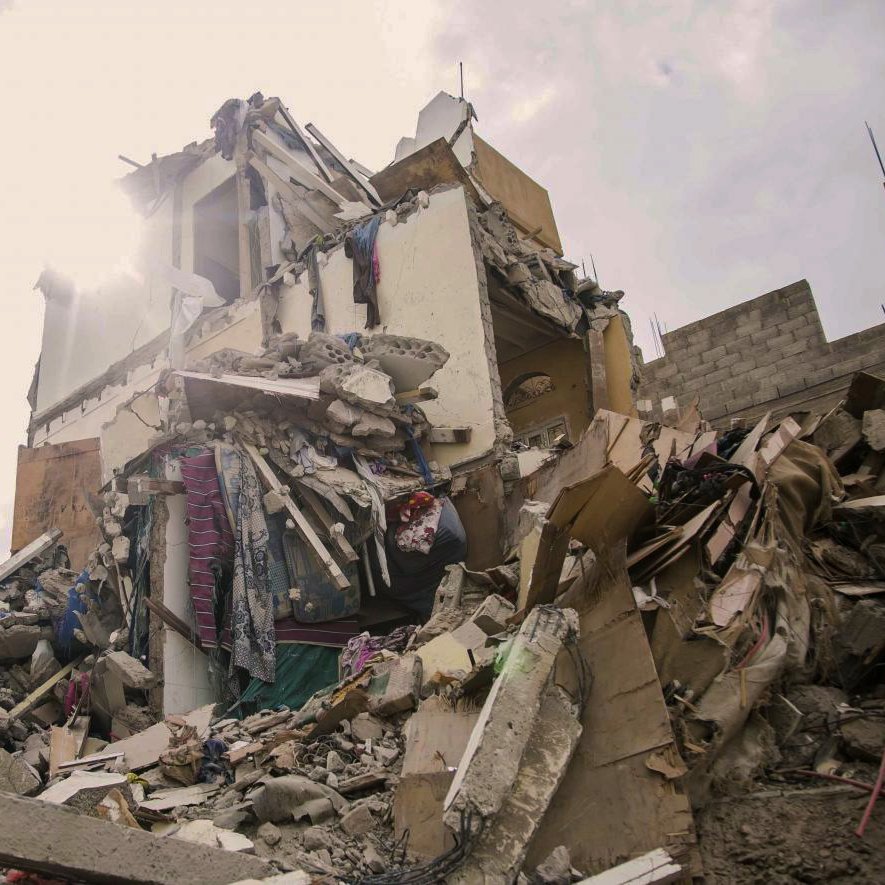 The image depicts a collapsed building, showing significant structural damage with debris scattered around. The upper portions of the structure are crumbled, and various materials like concrete, wood, and personal belongings are visible. In the foreground, there are a few individuals, likely responders or onlookers, inspecting the site. The scene is illuminated by sunlight breaking through the clouds, which adds a stark contrast to the devastation shown. The overall mood conveys a sense of disaster and the aftermath of a catastrophic event.
